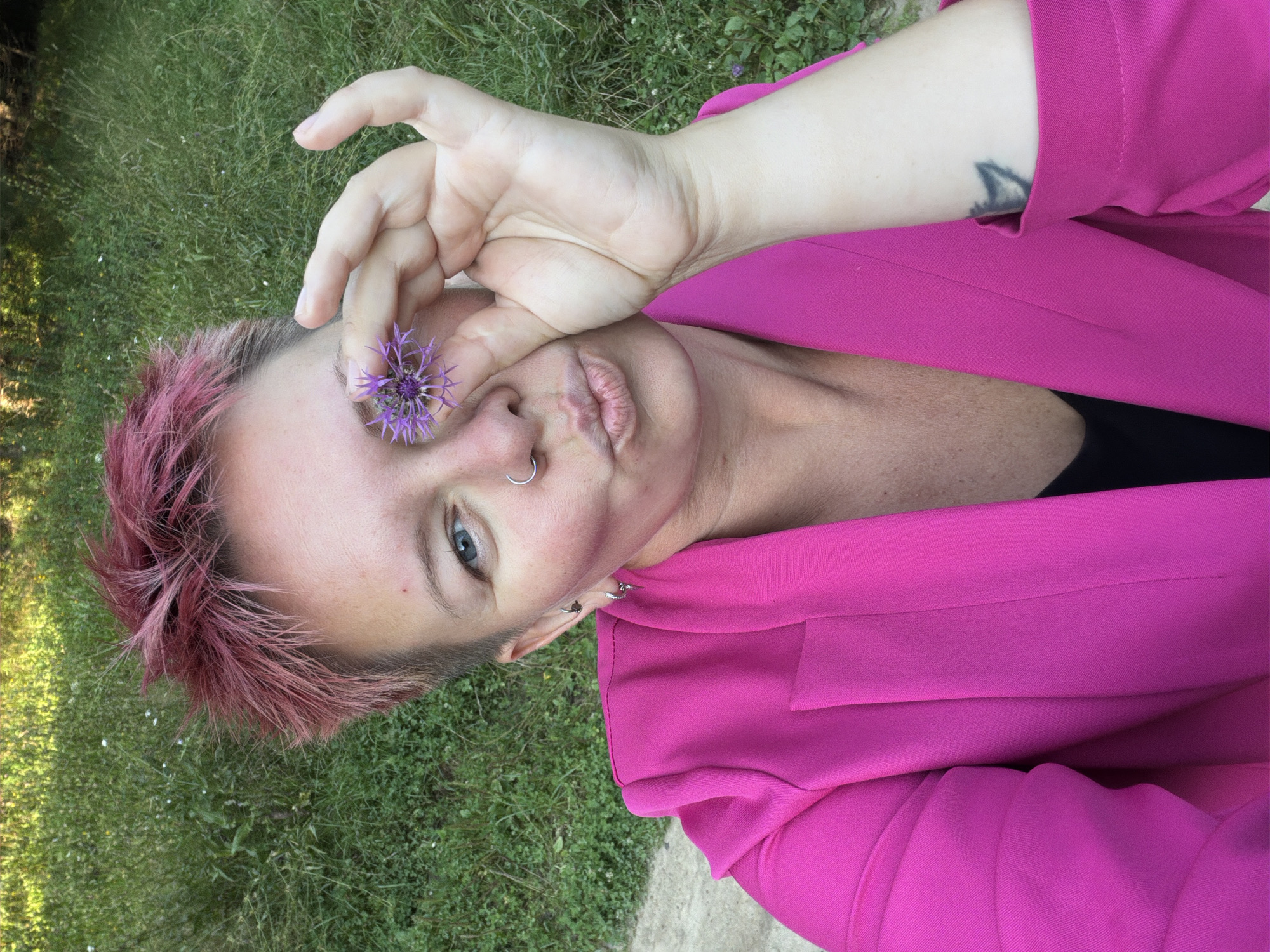 A woman with short, spiky pink hair and a nose ring is holding a small purple flower in front of her eye and making a kissing face. She is wearing a bright pink blazer over a black top. Her left arm is extended, and she has a tattoo on her forearm. The background is a grassy area with some sparse white flowers. The image appears to be a selfie taken outdoors, likely in a park or a natural setting. There is no specific information in the image to indicate the location in the city of Chaikovsky, Russia.