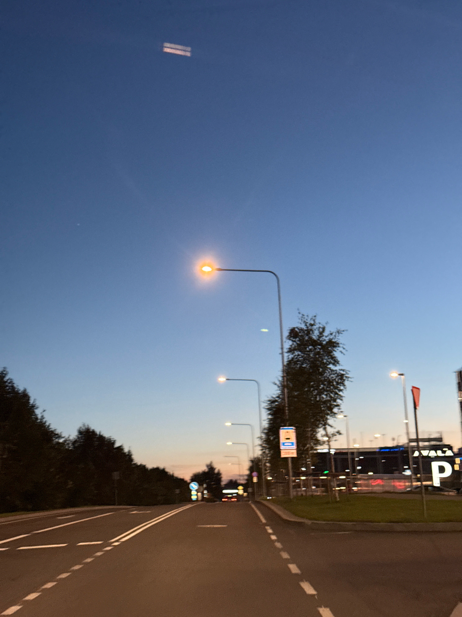 The image depicts an outdoor scene at dusk, likely in Tallinn, Estonia. A road with clear lane markings stretches into the distance, bordered by trees on the left and a developed area with buildings and streetlights on the right. Several streetlights are illuminated, casting a warm glow. In the background, a large building with illuminated signage, including the letter "P" and text partially obscured, is visible. The sky is a deep blue, transitioning to lighter hues near the horizon, with a subtle reflection or light streak visible high in the sky. The overall impression is of a suburban or edge-of-city location during the evening commute or early nightfall.