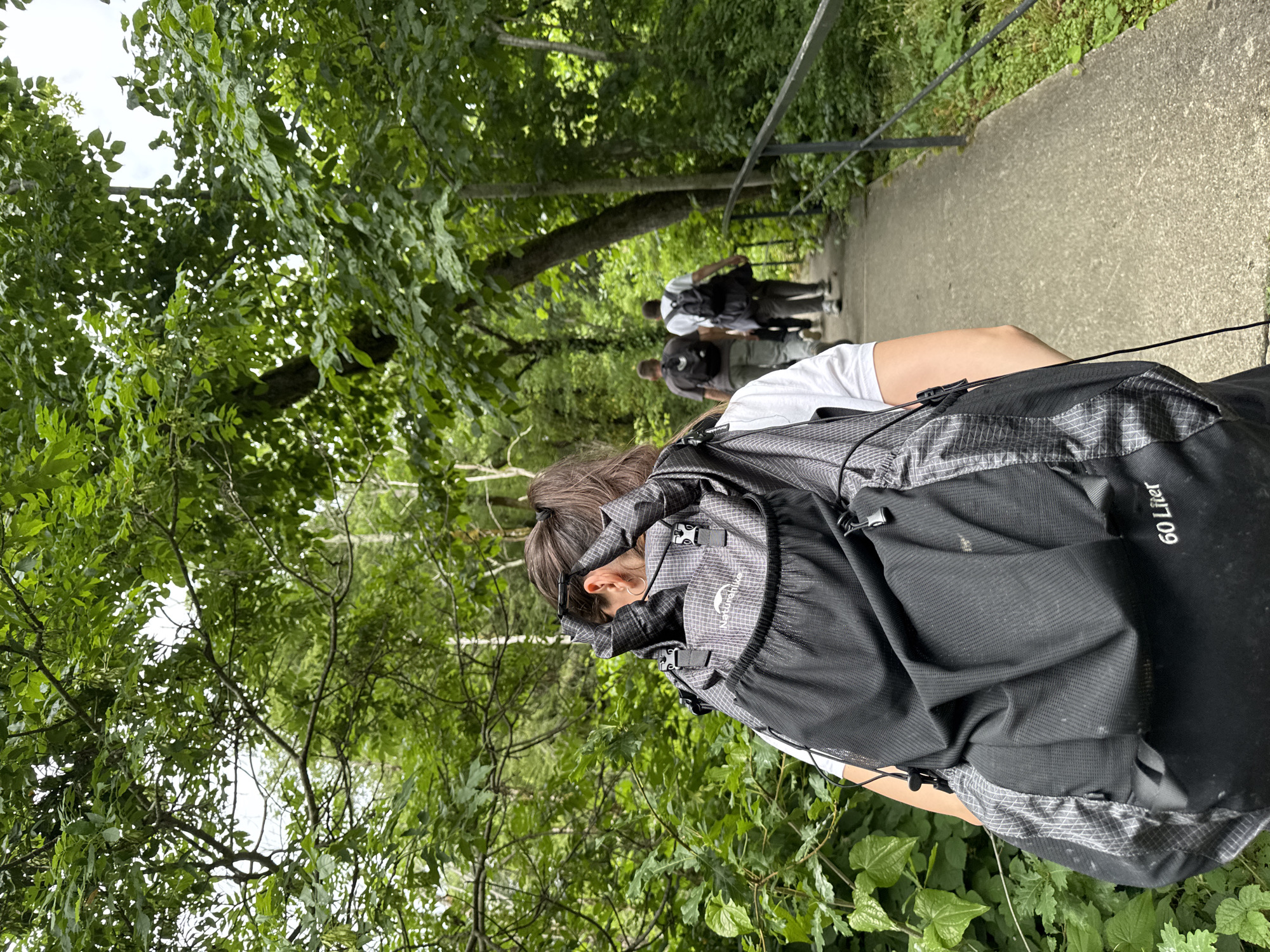 In Khleborob, Russia, a woman with dark hair pulled into a ponytail walks away from the camera on a paved path, wearing a large, black "60 Liter" backpack. She is dressed in a white t-shirt. Following her on the path are three men, also wearing backpacks and casual clothing. They are walking through a lush, green forest with dense foliage and trees lining the path, which is bordered by a black metal railing on the right side. The scene depicts a group hiking or walking in a natural, wooded area.