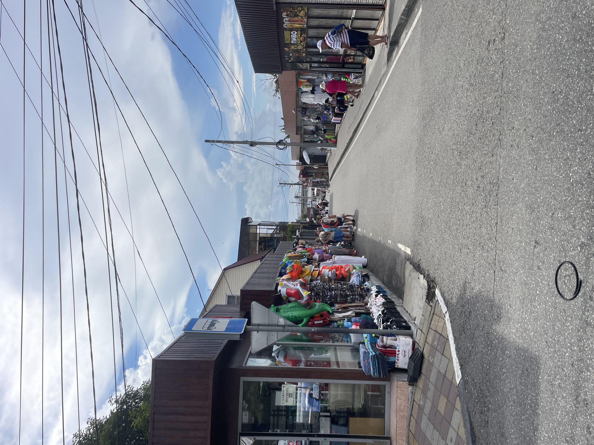 This image depicts a street scene in Sochi, Russia, likely a commercial area with numerous small shops and stalls lining the road. The street is paved and appears to be a one-way road, with shops on both sides. On the left side, there are several storefronts, one of which displays various beachwear and souvenirs, including inflatable toys like a large green crocodile. Some of these items are also displayed on the sidewalk. A bus stop sign is visible in front of one of the shops. Further down the street on the left, more shops are visible, with clothing and other goods displayed.
On the right side of the street, there are more shops, with one displaying a sign with images of beer and the word "ПИВО" (Pivo), suggesting it is a beer-selling establishment. People are seen walking along the sidewalks on both sides of the street, some browsing the shops, while others are walking. The sky is partly cloudy with blue skies visible. Overhead, a complex network of power lines crisscrosses the sky, supported by utility poles. The overall impression is of a bustling market street in a coastal city during daytime.