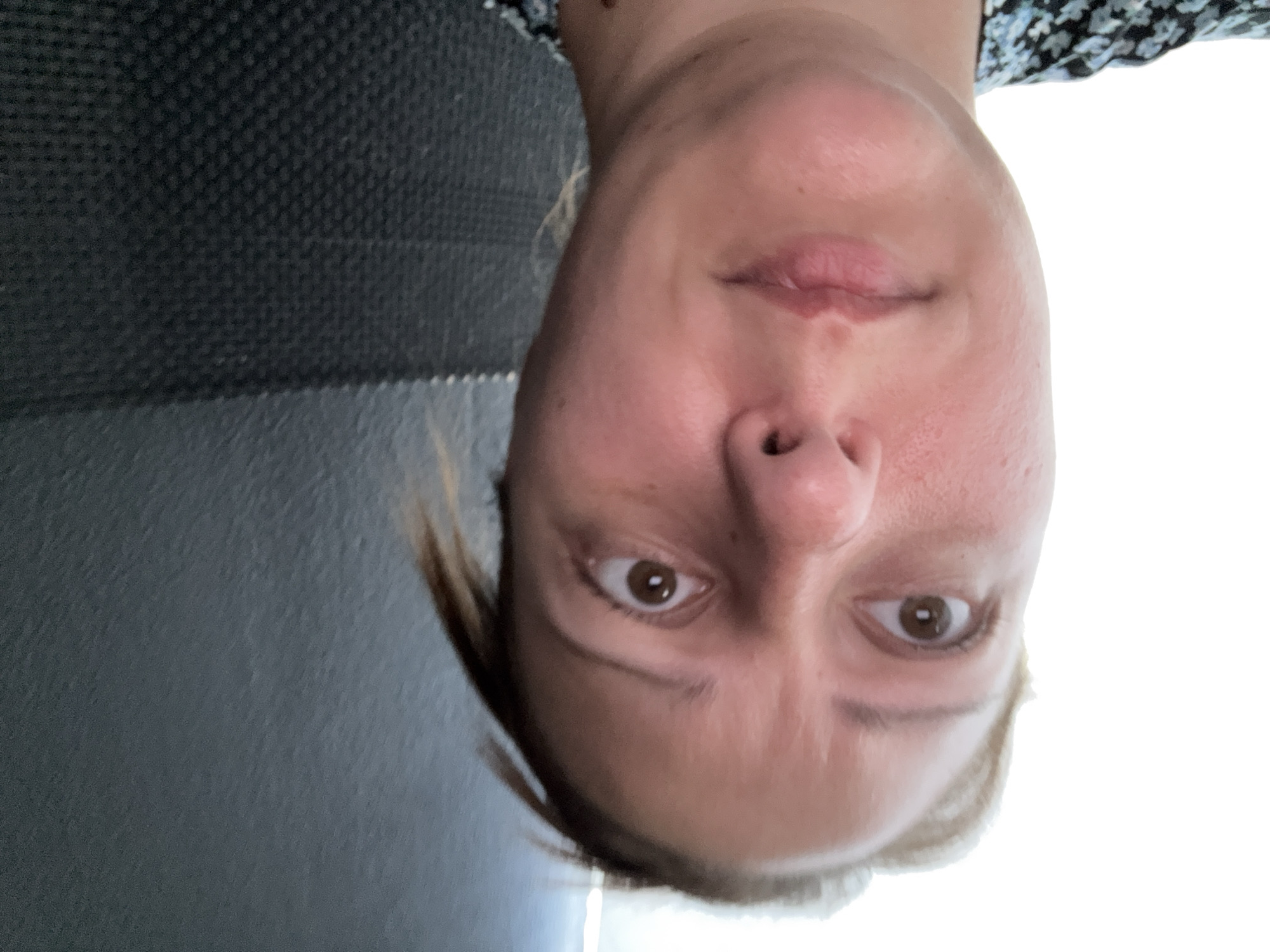 A medium shot, head-and-shoulders selfie shows a woman with light skin and blonde hair looking upwards with wide eyes and a contorted expression, suggesting surprise or fear. She is positioned in the left half of the frame, her face slightly angled to the right, with the background divided between a bright, undefined area on the left and a textured, dark gray wall on the right. Her eyes are brown and her eyebrows are furrowed. The lighting is harsh, with a strong light source coming from the left, casting shadows on the right side of her face. She is wearing a dark top with a floral pattern. The overall impression is one of an unguarded, candid moment captured by the camera.