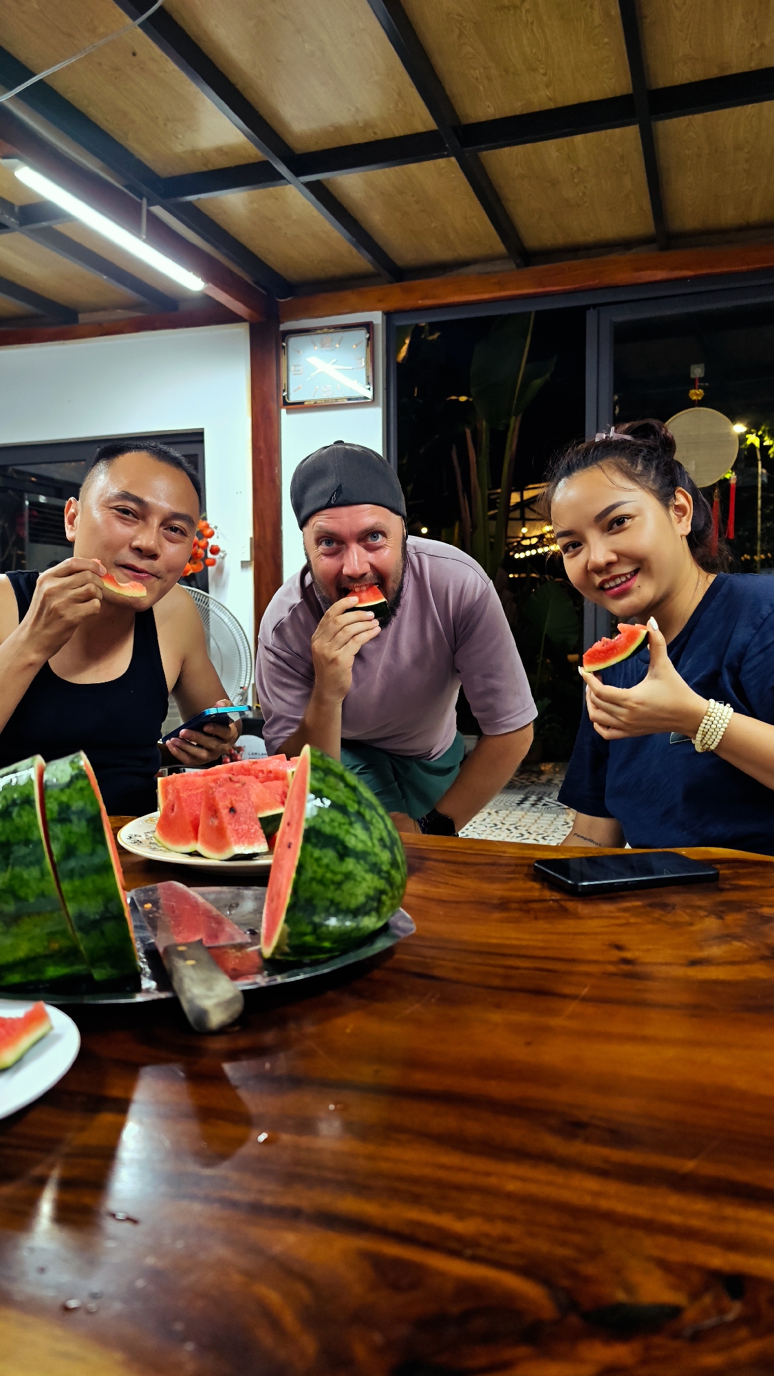 Three people are seated at a wooden table eating watermelon. A man on the left is wearing a black tank top, holding a phone. A man in the center is wearing a black hat and a light purple shirt. A woman on the right is wearing a dark blue shirt and a white beaded bracelet. A large watermelon, partially sliced, sits on a silver platter along with a knife. Watermelon slices are on a white plate. Cell phones are on the table. The location is in the city of Hà Giang 1 Ward, Vietnam. A clock is on the wall in the background. Tropical plants are visible through a window.