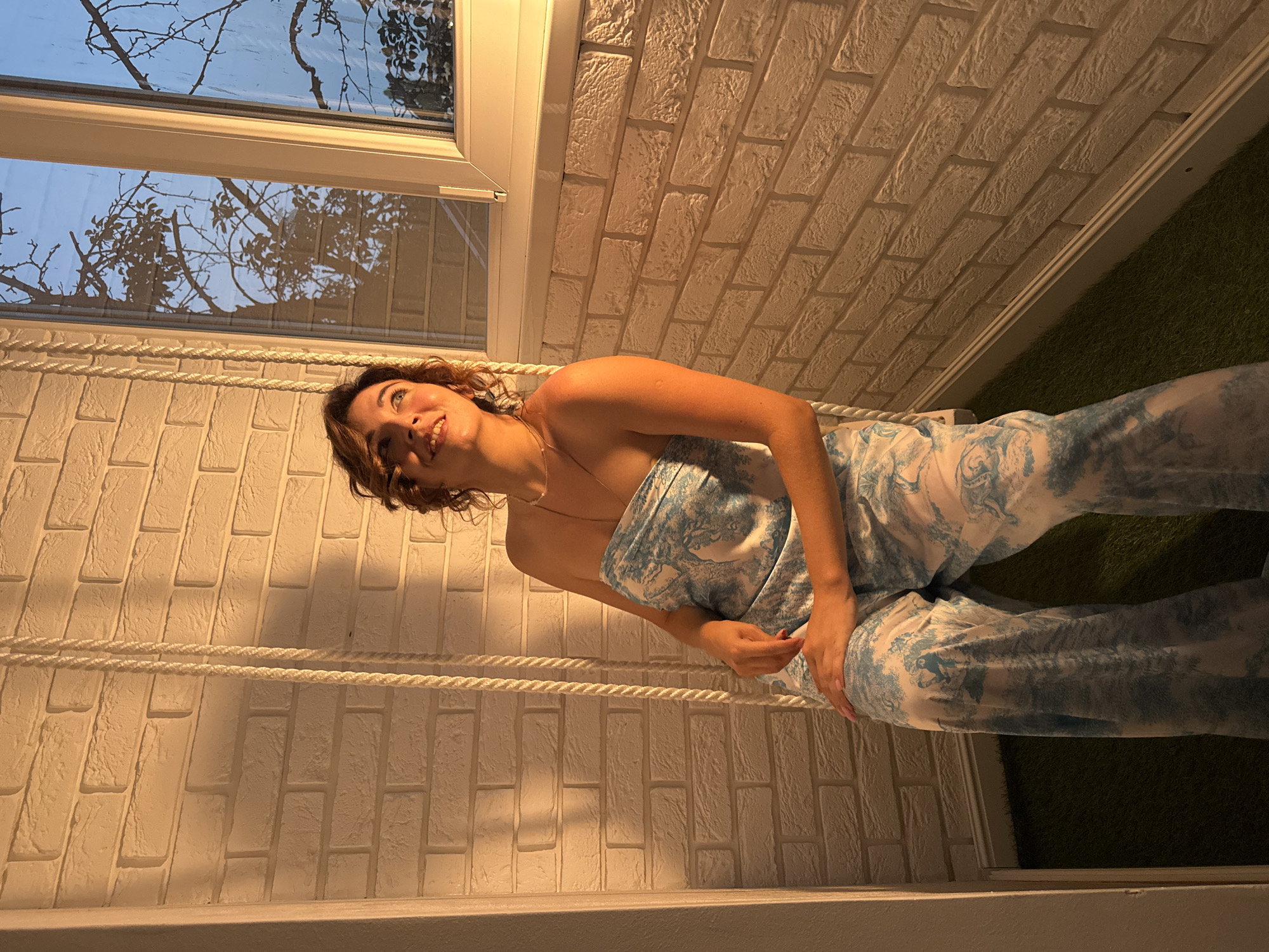 A young woman with brown hair is lying on a swing, looking up and smiling. She is wearing a white and blue patterned outfit. The swing is suspended by thick white ropes. Behind her, a wall is decorated with white bricks, and a window shows a silhouette of tree branches against a pale sky, suggesting it is either dawn or dusk. To the right of the brick wall, a patch of green artificial turf is visible. The lighting in the scene is warm and appears to be artificial, casting shadows on the brick wall. The overall mood is relaxed and cheerful. No text is visible in the image.