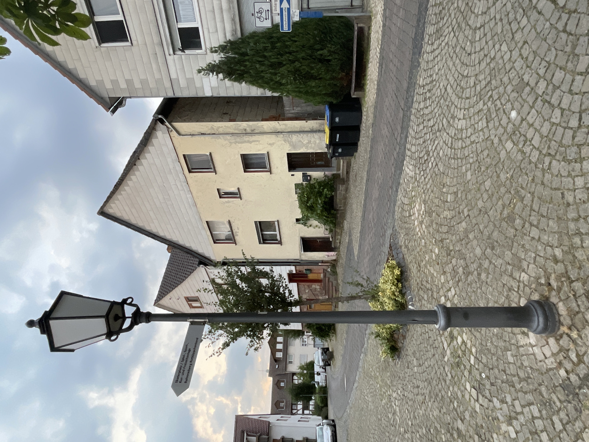 In Neukirchen, Germany, a gray streetlamp stands in the foreground on a cobblestone street. A directional signpost is attached to the lamp post, pointing left and indicating "Skaterplatz / Spiel-, Sport-, Kur- und Freizeitanlage Bahnradweg Rotkäppchenland". Behind the lamp post, two houses are visible. The house on the left is a light-colored, two-story building with a triangular roof. The house on the right is a taller, light-colored building with a flat roof, and a green evergreen tree is planted next to it. The sky is cloudy.