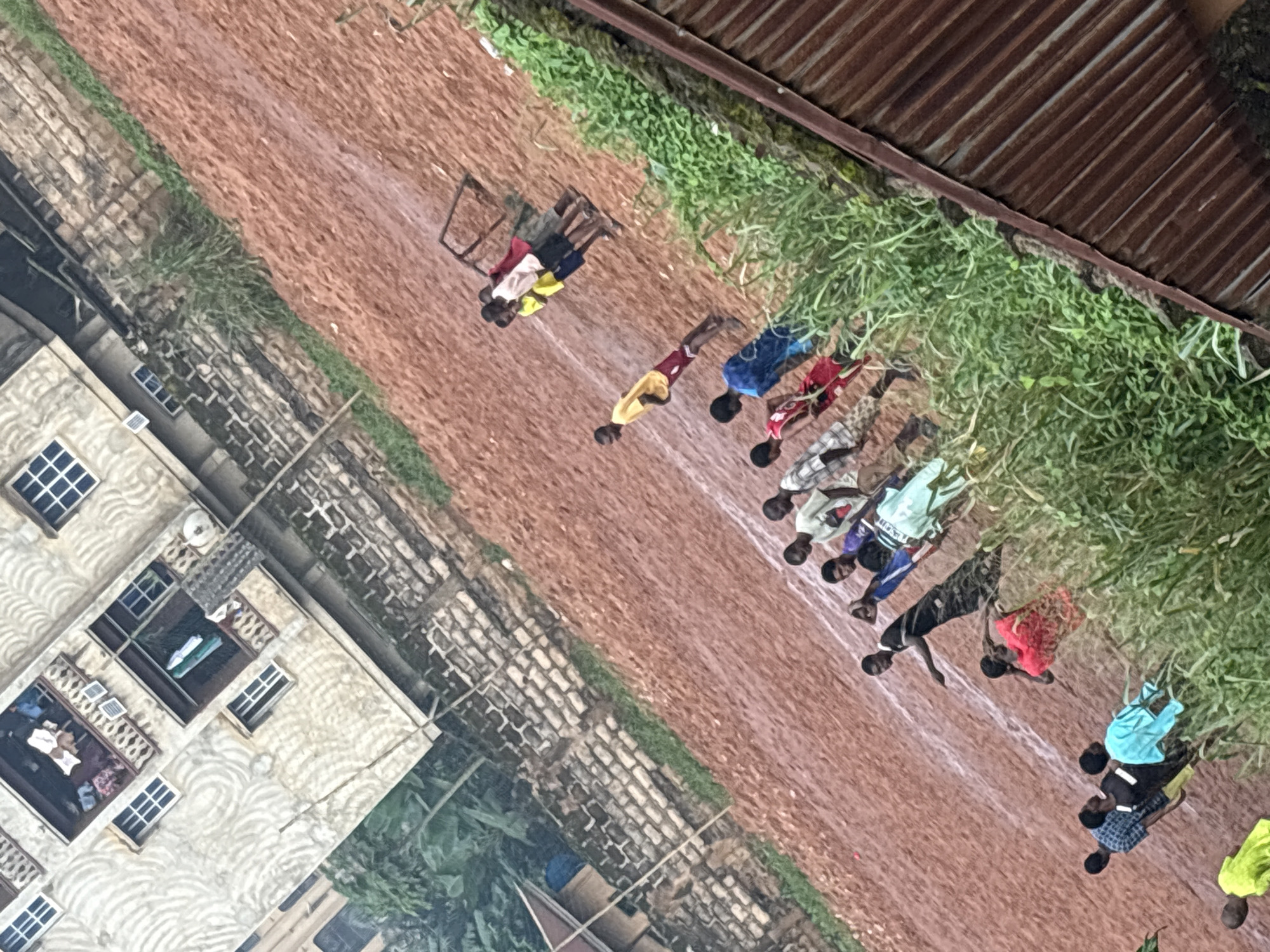 The image depicts an outdoor scene in a densely populated area of Onitsha, Nigeria. In the foreground, a group of young people, primarily males, are gathered on a dirt field. Some appear to be participating in a sporting activity, as evidenced by the lines drawn on the field, suggesting a track or court. The field is reddish-brown and appears wet, possibly from rain.
Several individuals are visible in various states of engagement. One person in a yellow shirt and red shorts stands near the top of the frame, facing away from the camera. Closer to the bottom, a group of individuals are lined up, with some wearing brightly colored clothing like blue and red. A person in a black and white checkered shirt is also present.
In the background, a multi-story building with balconies and windows is visible. The architecture suggests it is an urban dwelling common in Nigeria. Adjacent to the building is a wall constructed of bricks. Lush green vegetation, including tall grass and possibly some trees, borders the field and the building. A corrugated metal roof of a structure is visible on the far right of the image. The overall impression is of a vibrant, possibly informal, community gathering or event taking place in a characteristic urban Nigerian setting.