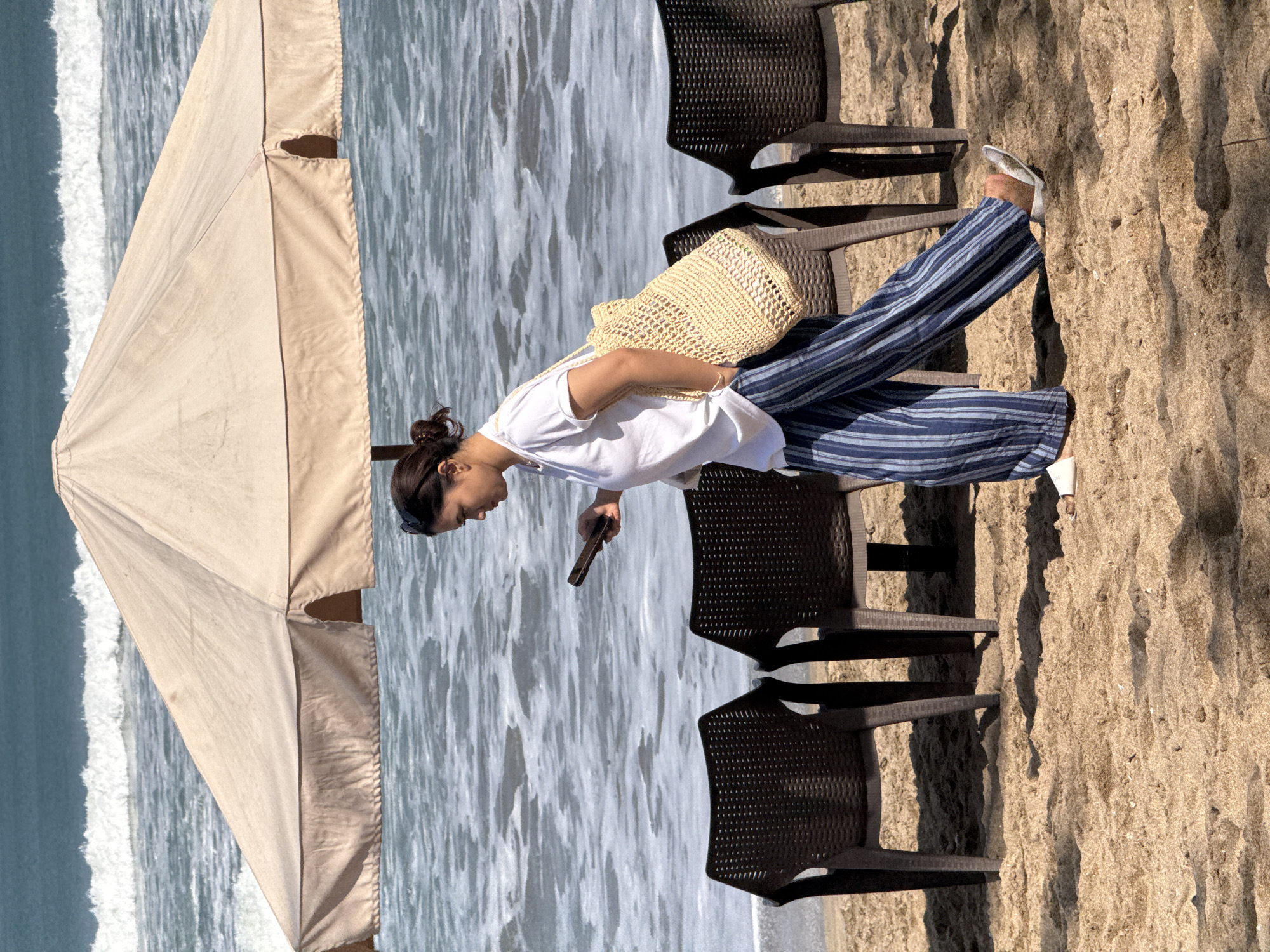 A woman with her hair pulled back in a bun walks on a sandy beach in Kuta, Indonesia, holding a smartphone and carrying a straw bag over her shoulder. She is wearing a white t-shirt and blue and white striped pants. The background shows the ocean with waves and a large, beige umbrella providing shade. Several dark plastic chairs are arranged on the sand.