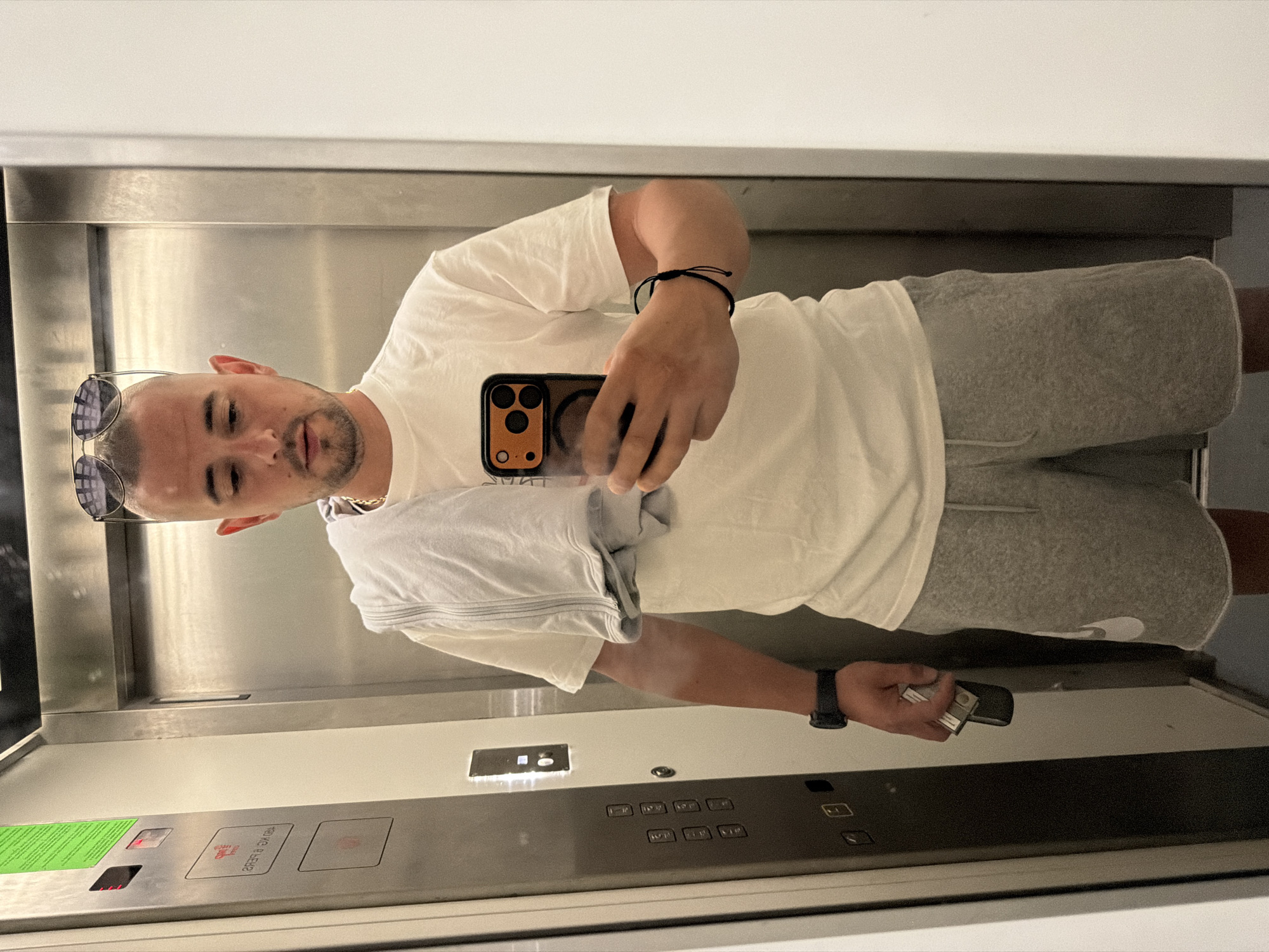 A young man with a shaved head and sunglasses on top of his head takes a mirror selfie in an elevator. He is wearing a white t-shirt, grey athletic shorts, and a black wristwatch. A light blue towel is draped over his left shoulder. In his right hand, he holds a smartphone with an orange case, and in his left hand, he holds a silver object. The elevator has stainless steel doors and a panel with buttons and a green sign. The location is an elevator in Burgas, Bulgaria.
