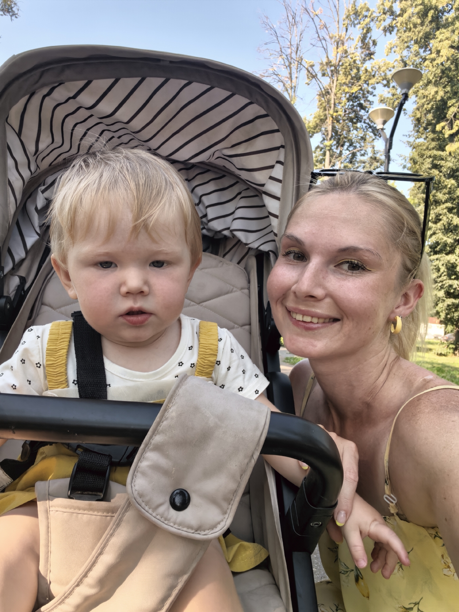In a sunny park setting, a blonde woman and a young child are in a stroller. The woman, smiling and looking at the camera, wears a yellow dress and yellow hoop earrings. The child, with blonde hair, sits in the stroller wearing a white shirt with a black and white floral pattern and yellow suspenders. The stroller has a striped interior lining. Trees and a lamppost are visible in the background, suggesting an outdoor urban environment. The image does not provide any specific details to indicate the location is Sterlitamak, Russia.