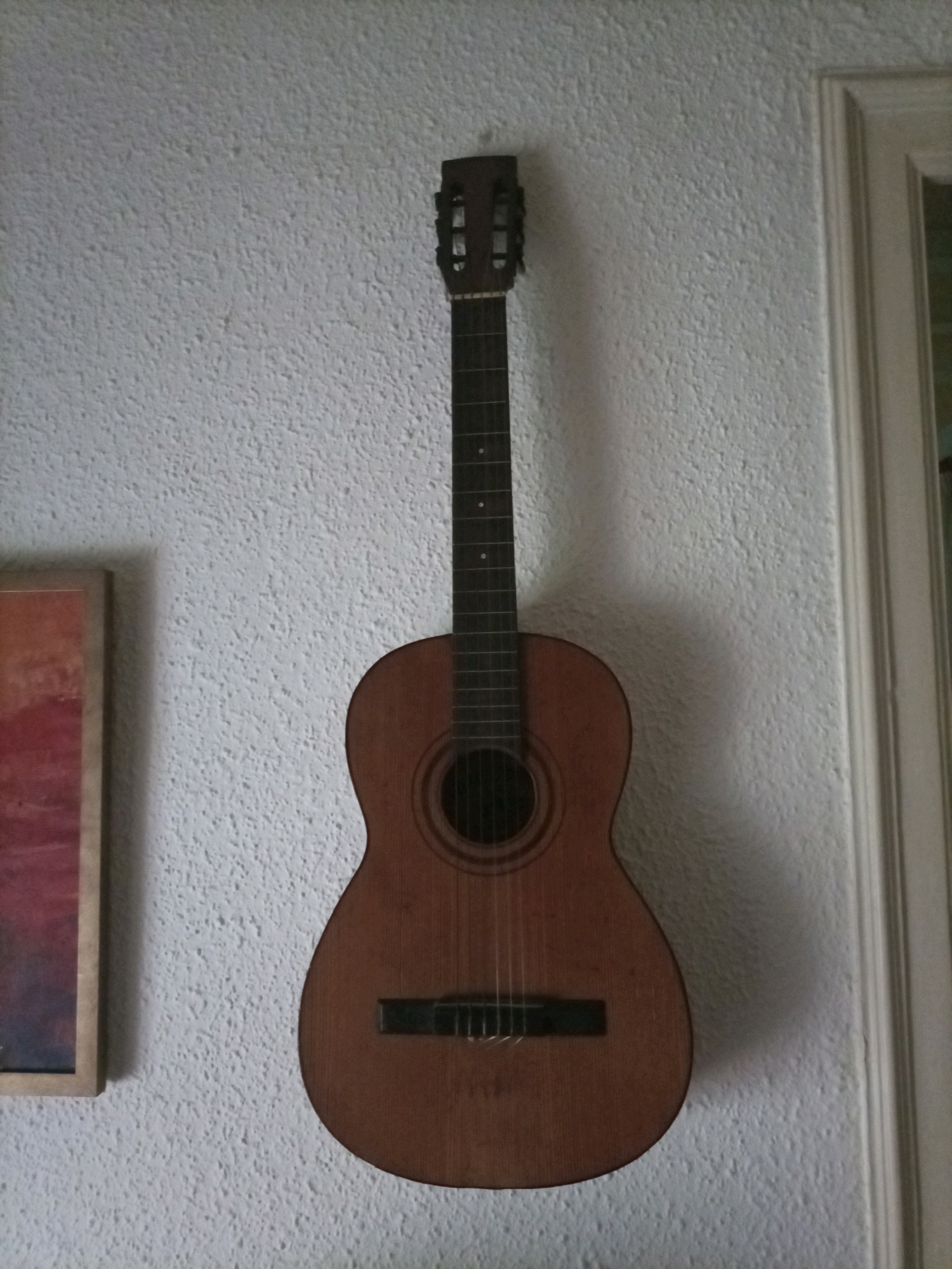 An acoustic guitar is hanging on a textured white wall. To the left of the guitar is a framed picture with a colorful abstract painting. To the right of the guitar, a portion of a door frame is visible. The lighting suggests an indoor setting. The scene does not provide enough information to determine its location in Tarragona, Spain.