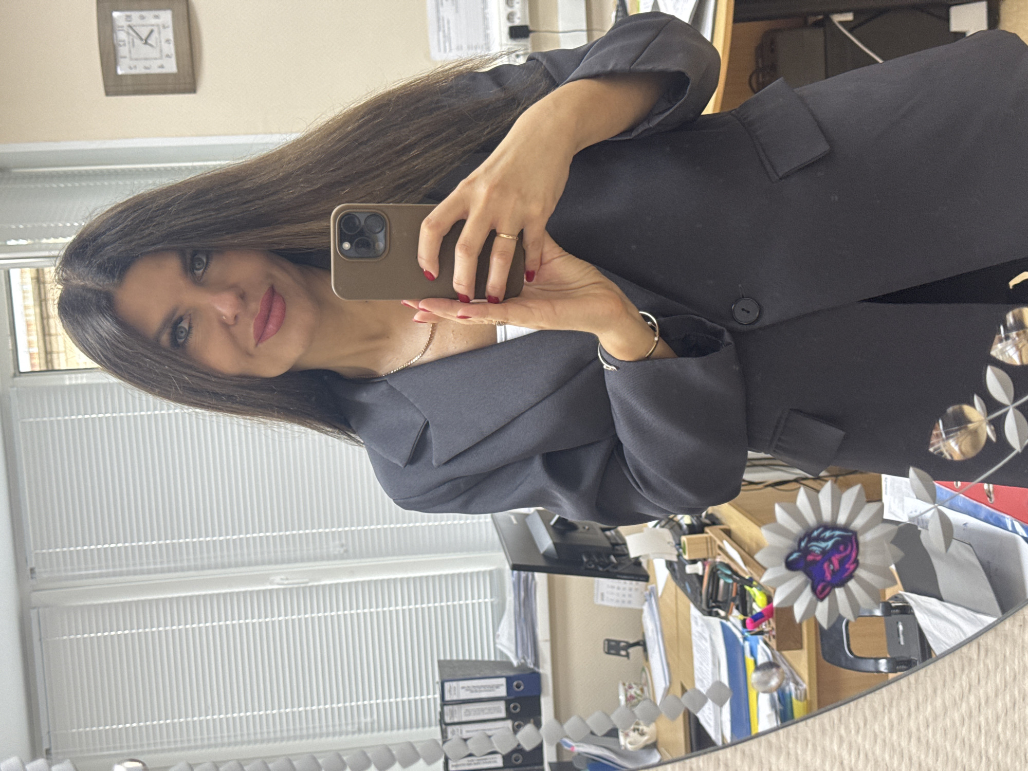 In a reflection in a mirror, a woman with long dark hair and blue eyes is taking a selfie. She is wearing a dark blazer over a white top and has red nail polish. She is holding a smartphone in her right hand and wearing a gold ring on her left ring finger and a silver bracelet on her left wrist. The background appears to be an office setting, with blinds on a window, a computer monitor, stacks of papers, folders, and a clock on the wall. The overall impression is a professional or office environment, possibly in the city of Chaikovsky, Russia.