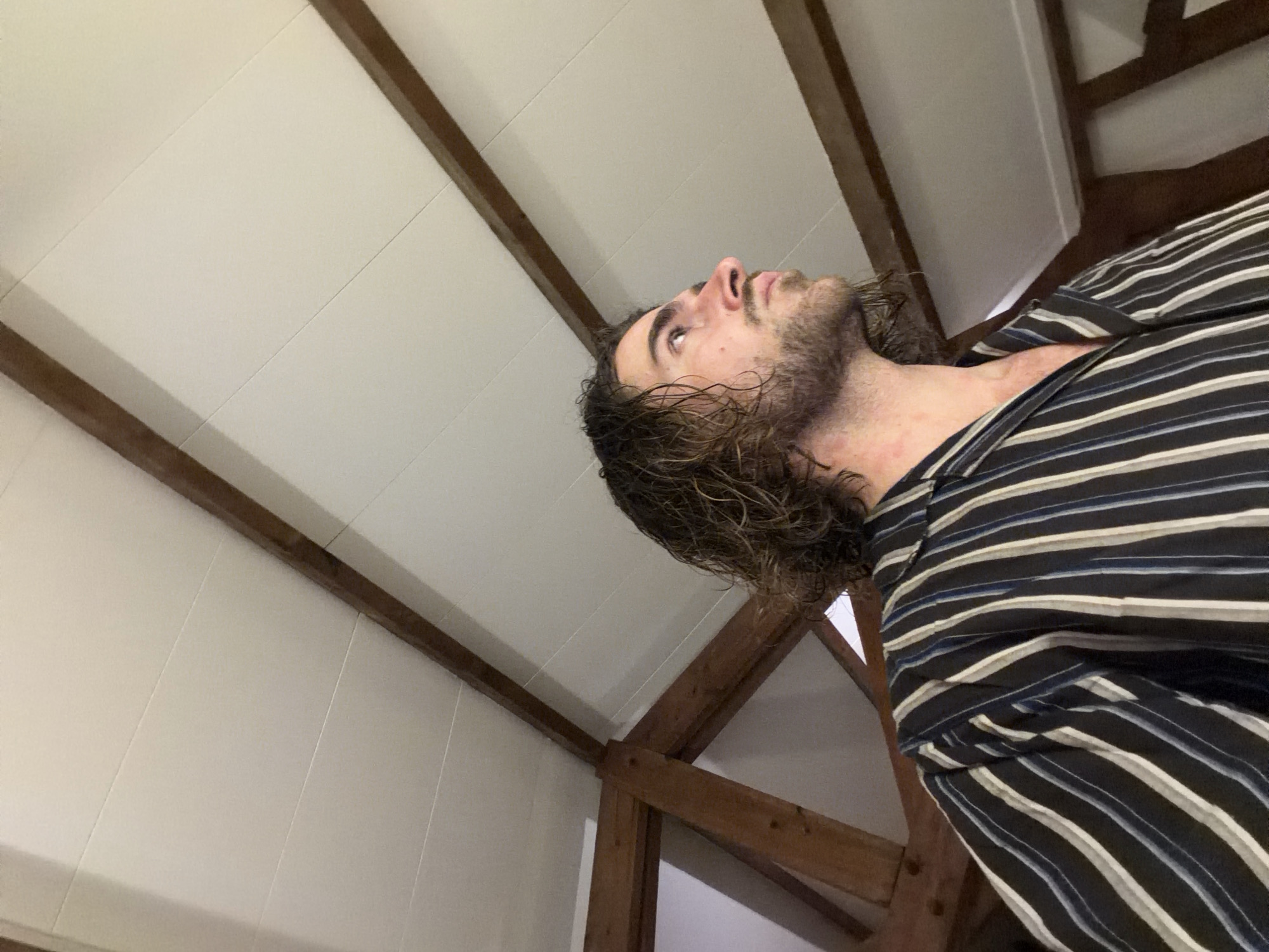 A low-angle shot depicts a man with long, wavy brown hair and a beard, wearing a striped shirt. He is looking upwards. The background reveals an interior space with white paneled walls and wooden beams suggesting an attic or loft conversion. The location is not identifiable as Paris, France based on the visual information provided.