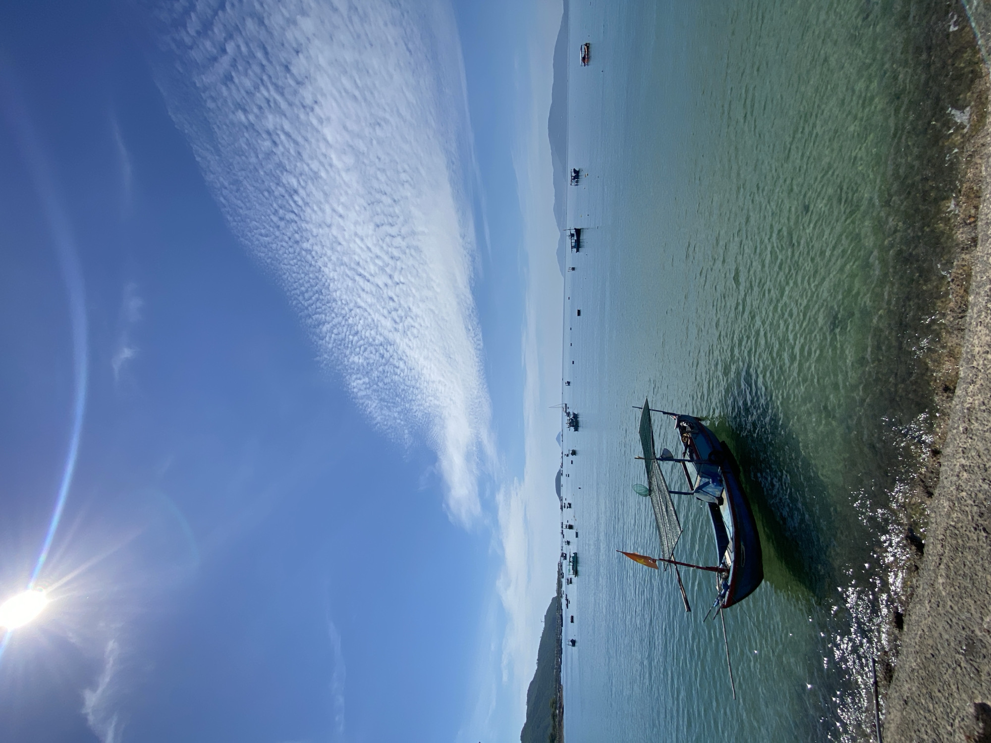 The image depicts a scenic view of the sea near Nha Trang, Vietnam, under a clear blue sky with scattered, textured clouds. The sun is prominently visible in the upper left corner, casting bright light and a rainbow-like lens flare. In the foreground, a small, blue fishing boat with a canopy and an orange flag is anchored in the shallow, turquoise water. The water is clear enough to reveal the rocky seabed. A concrete embankment is visible in the lower left corner, with some debris. The middle ground and background show a calm sea dotted with numerous other fishing boats of various sizes. In the distance, a coastline with rolling hills and mountains is visible, suggesting the proximity to land. The overall atmosphere is serene and peaceful, capturing a typical day in a coastal Vietnamese fishing village.