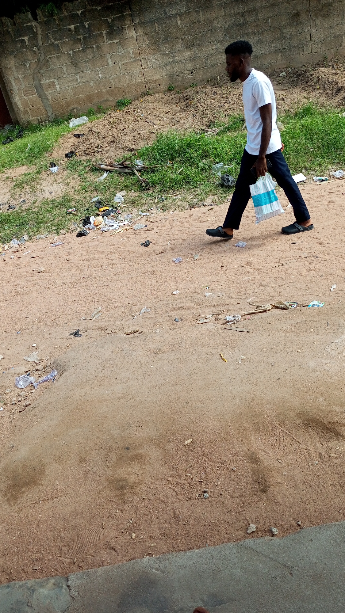 A young Black man wearing a white t-shirt and black pants walks across a sandy, debris-strewn area. He carries a white and blue striped shopping bag in his right hand and a black mobile phone in his left. The man is seen from the side, walking towards the right of the frame. The ground is covered with sand, grass, and various types of litter, including plastic bags, wrappers, and other refuse. In the background, a low, grey concrete block wall runs horizontally across the image, with patches of green vegetation growing in front of it. The overall scene suggests an outdoor environment, possibly on the outskirts of a city or in a neglected urban area.