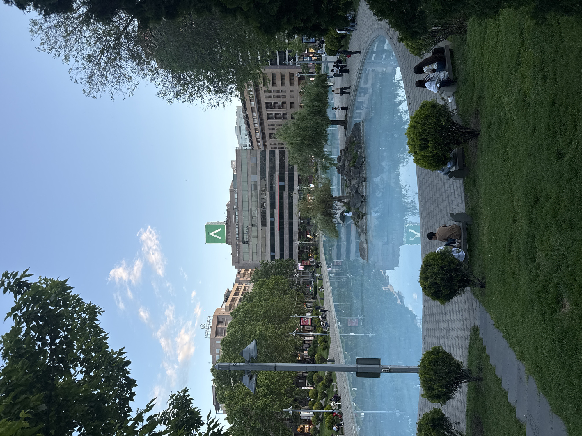 The image depicts a scene in Yerevan, Armenia. The viewpoint is looking across a long, narrow rectangular water feature that serves as a reflecting pool, displaying the reflection of buildings, trees, and the sky. On either side of the pool are walkways paved with interlocking bricks or stones. People are sitting on benches along the walkways. The buildings are a mix of modern and older architecture. The foreground features trees and well-maintained grassy areas with trimmed shrubbery. A V-shaped logo is prominently displayed on a tall building in the background. Streetlights and signage are visible along the walkway. The sky is blue with scattered clouds.