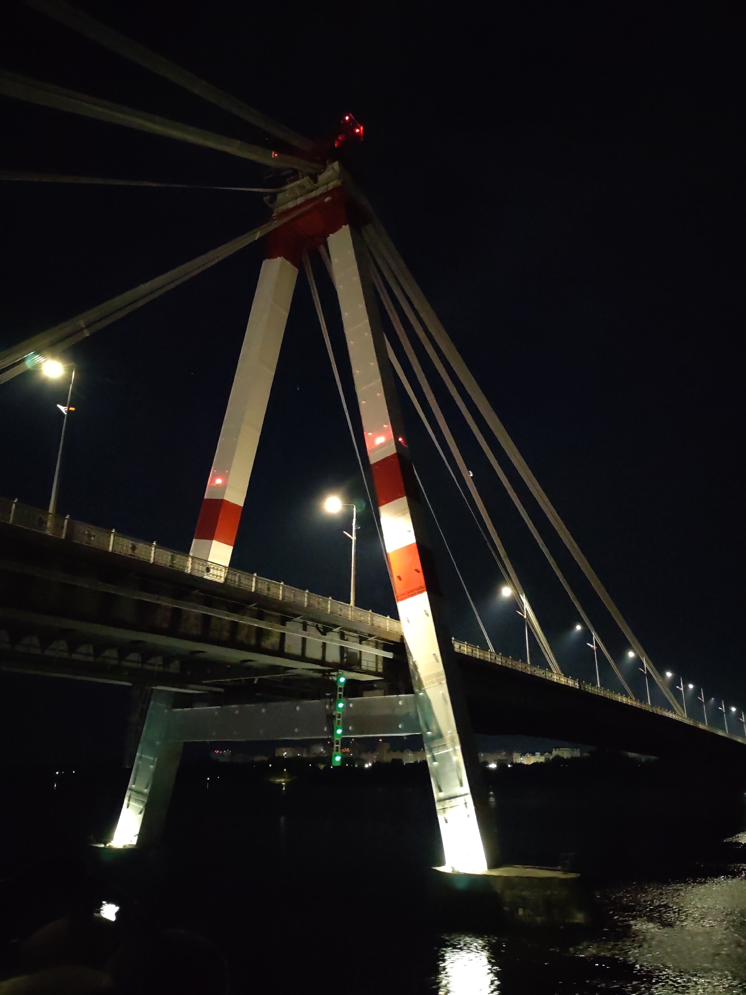 This nighttime image depicts the North Bridge in Kyiv, Ukraine. The bridge is a cable-stayed structure, and its towers are painted with alternating red and white bands. Cables extend from the top of the towers, supporting the bridge deck. Streetlights illuminate the roadway. The bridge spans a body of water, and reflections of light sources are visible on the surface. The sky is dark, and minimal details can be discerned in the background cityscape.