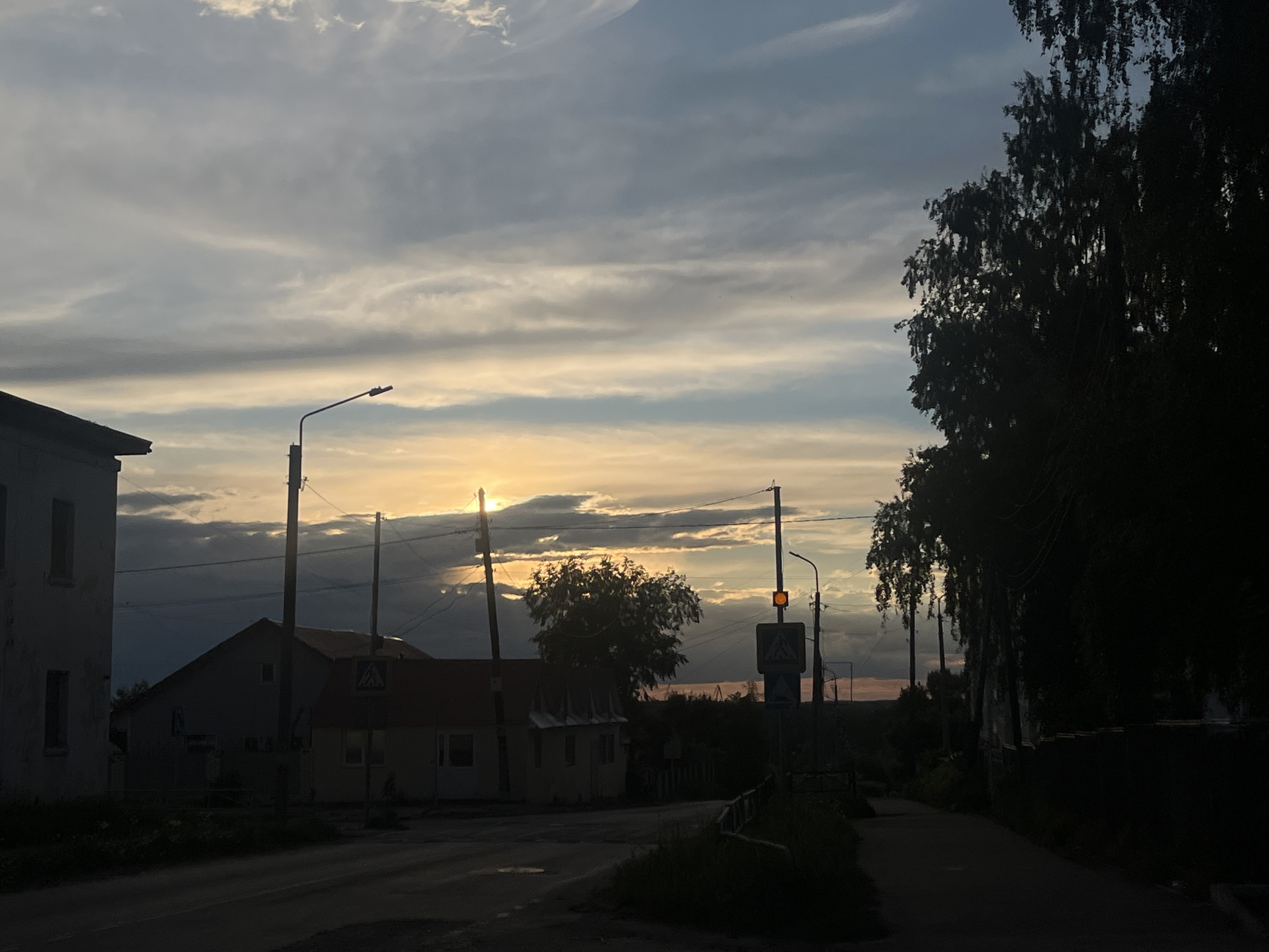 The image depicts a street scene in the city of Chaikovsky, Russia, at dusk or dawn. On the left, there is a two-story building with visible windows. A one-story building with a red roof stands adjacent to the first building. A street lamp and a pedestrian crossing sign are visible nearby. Wires stretch across the sky, intersecting with the lampposts. A tree stands in the middle ground, partially obscuring the horizon. On the right side of the image, a row of trees casts shadows on the road. The road itself is dimly lit. The sky is filled with clouds, with a faint glow of sunlight breaking through.