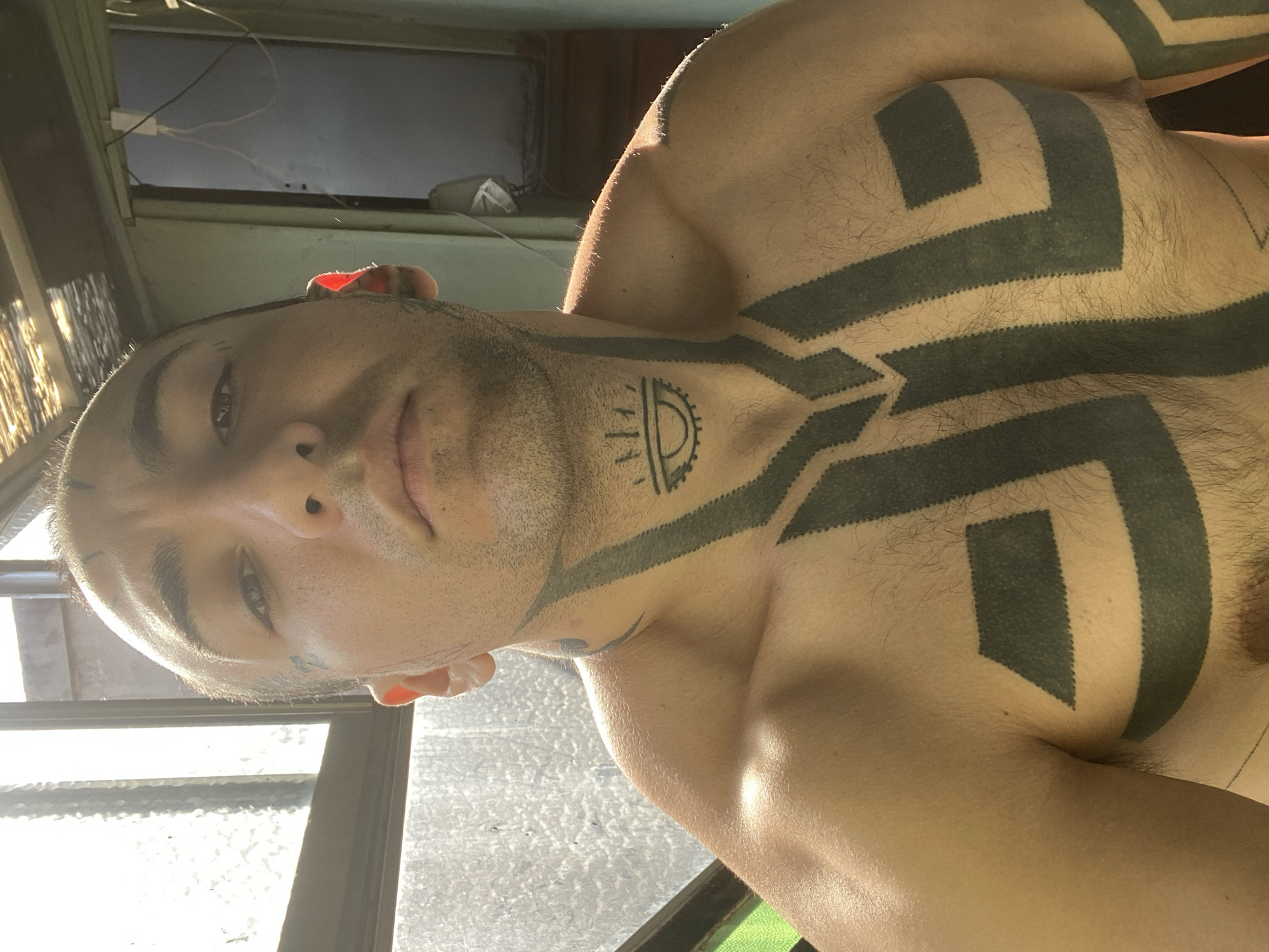 A close-up, medium-shot photograph depicts a light-skinned male with short, light-colored hair, stubble, and prominent dark tattoos across his chest and neck. The subject is positioned at an angle, looking downwards towards the camera, with a slight shadow across his face. The tattoos are tribal-style, consisting of thick, black geometric shapes. A window with frosted glass is visible in the background, through which some sunlight filters. A pale green surface is partially visible beneath the window. The location cannot be determined from the image.