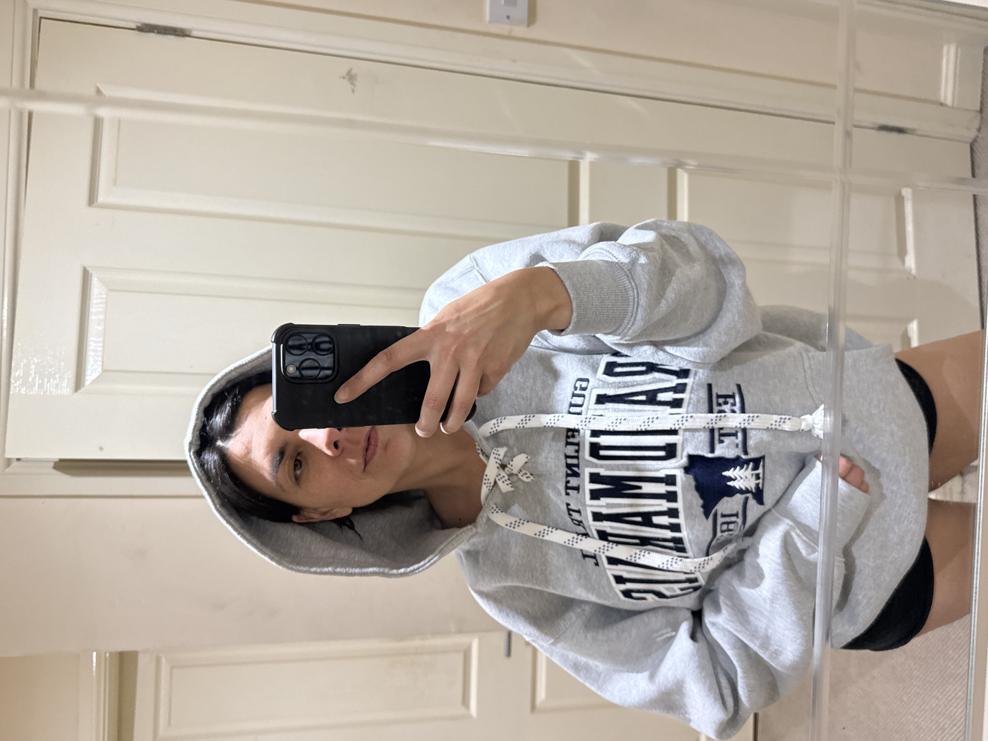 A person is taking a mirror selfie in what appears to be a domestic setting. The person is wearing a grey hooded sweatshirt with white laces and dark lettering on the front, along with dark shorts. They are holding a black smartphone up to take the picture, partially obscuring their face. The background shows a cream-colored door with paneling. There is no information in the image that indicates the location is in the city of Great Yarmouth, United Kingdom.