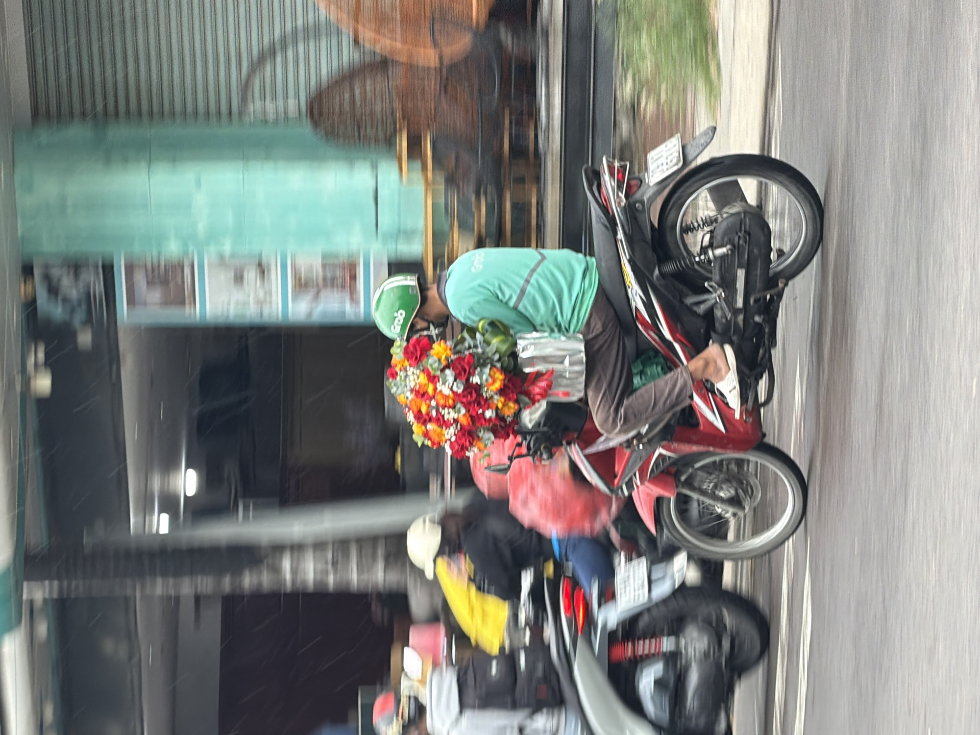 The image depicts a street scene in Nha Trang, Vietnam, with motion blur indicating movement. The primary subject is a person riding a red motorcycle, wearing a green shirt with "Grab" logo, a green helmet with a "Grab" logo, and brown pants. They are carrying a large bouquet of red, orange, and yellow flowers. To the left, another motorcycle with a rider in a grey hoodie and backpack is partially visible, as is a person wearing a yellow jacket. The background shows buildings with metal shutters and what appear to be shops. The street is wet, suggesting recent rain, and there are white lines painted on the road.