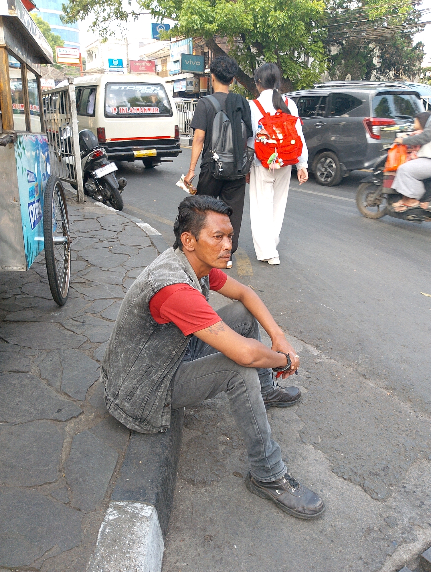 In Bandung City, Indonesia, a man sits on the curb of a street, his legs crossed and his hands clasped. He wears a gray denim vest over a red t-shirt, gray pants, and dark work boots. To his left, a motorcycle is parked next to a food cart. Behind the man, two young people walk away from the camera. The person in front, wearing a backpack, is partially obscured by a woman with a red backpack. In the background, a white van with "STASIUN LOKAL" written on its rear is visible. Cars and trees line the street, with buildings and utility poles in the distance. The scene appears to be a typical street view in an urban environment.