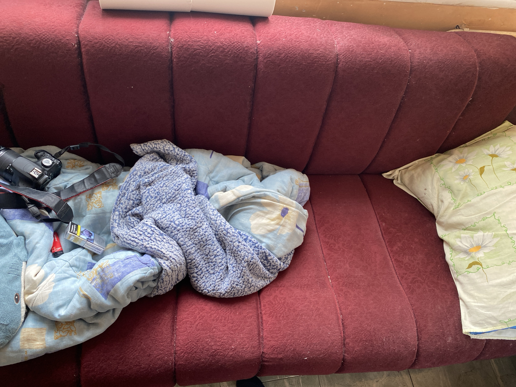 A dark red sofa is heavily cluttered. From left to right, there is a camera with a lens and strap, followed by a blue patterned blanket with a stuffed shark on it, and a pack of cigarettes. A blue knitted blanket is crumpled in the center. On the right side of the sofa is a white pillow with green flower patterns. The sofa has a patterned fabric and the room seems to be indoors. I am unable to determine the location of Sofia, Bulgaria from the image.