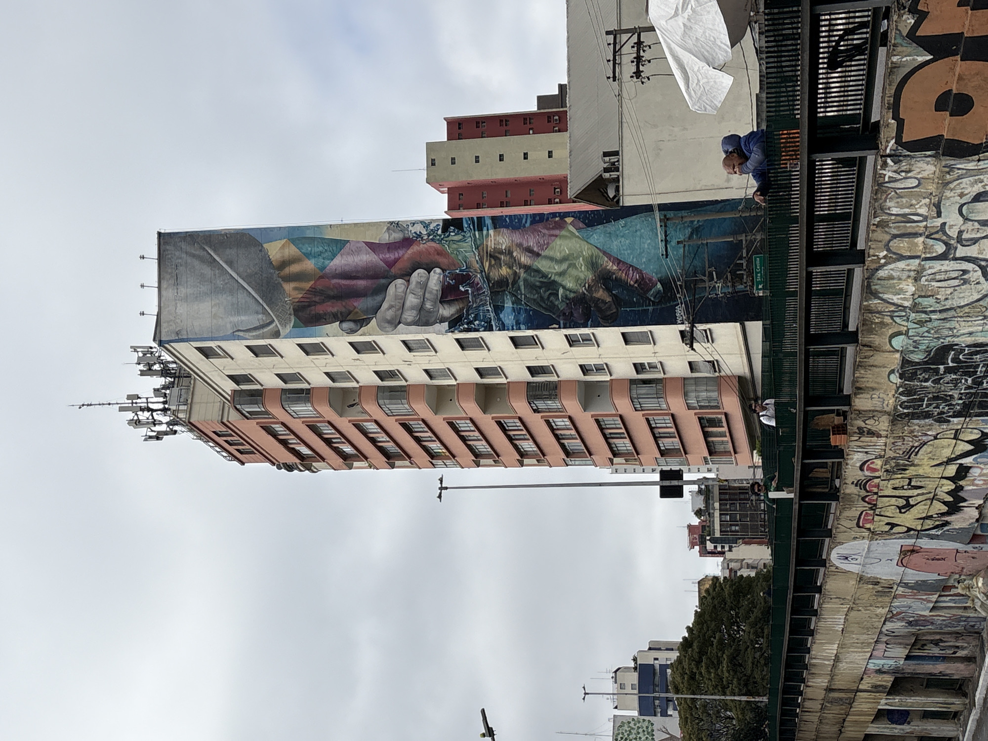 The image depicts a street scene in the city of São Paulo, Brazil. A tall, multi-story building with a pink facade and numerous windows dominates the center of the frame. The building's side is adorned with a large, vibrant mural featuring two hands reaching for each other, with water splashing between them and abstract geometric shapes in various colors.
In the foreground, a bridge or elevated walkway is visible, lined with a dark green railing. Graffiti covers the lower walls of this structure. Several people are present on the walkway, including a man in a blue hooded jacket and orange pants who is leaning on the railing and appears to be covering his mouth with his hand. Another person, wearing a white shirt and glasses, stands further down the walkway.
Behind the main building, other structures of varying heights and colors, including a red and beige building, are visible. The sky is overcast. A street lamp and electrical wires are also present in the scene. The location is identified as being in São Paulo, Brazil, specifically on Rua Thaumaturgo de Azevedo, as indicated by a green street sign on the mural.