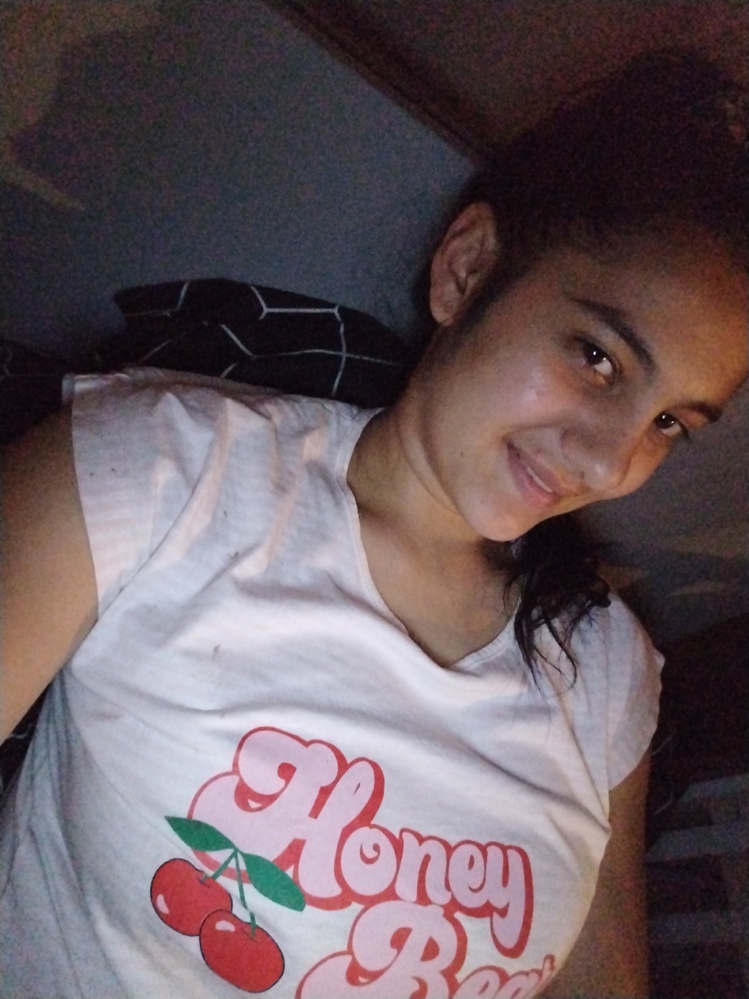 A close-up, overhead shot shows a young woman with dark hair pulled back in a ponytail, lying down and looking towards the camera. She is wearing a white t-shirt with a graphic of two red cherries and the words "Honey Bear" printed in pink. The background is dark and out of focus, with hints of a patterned pillow behind her. The lighting is dim, suggesting an indoor setting at night.