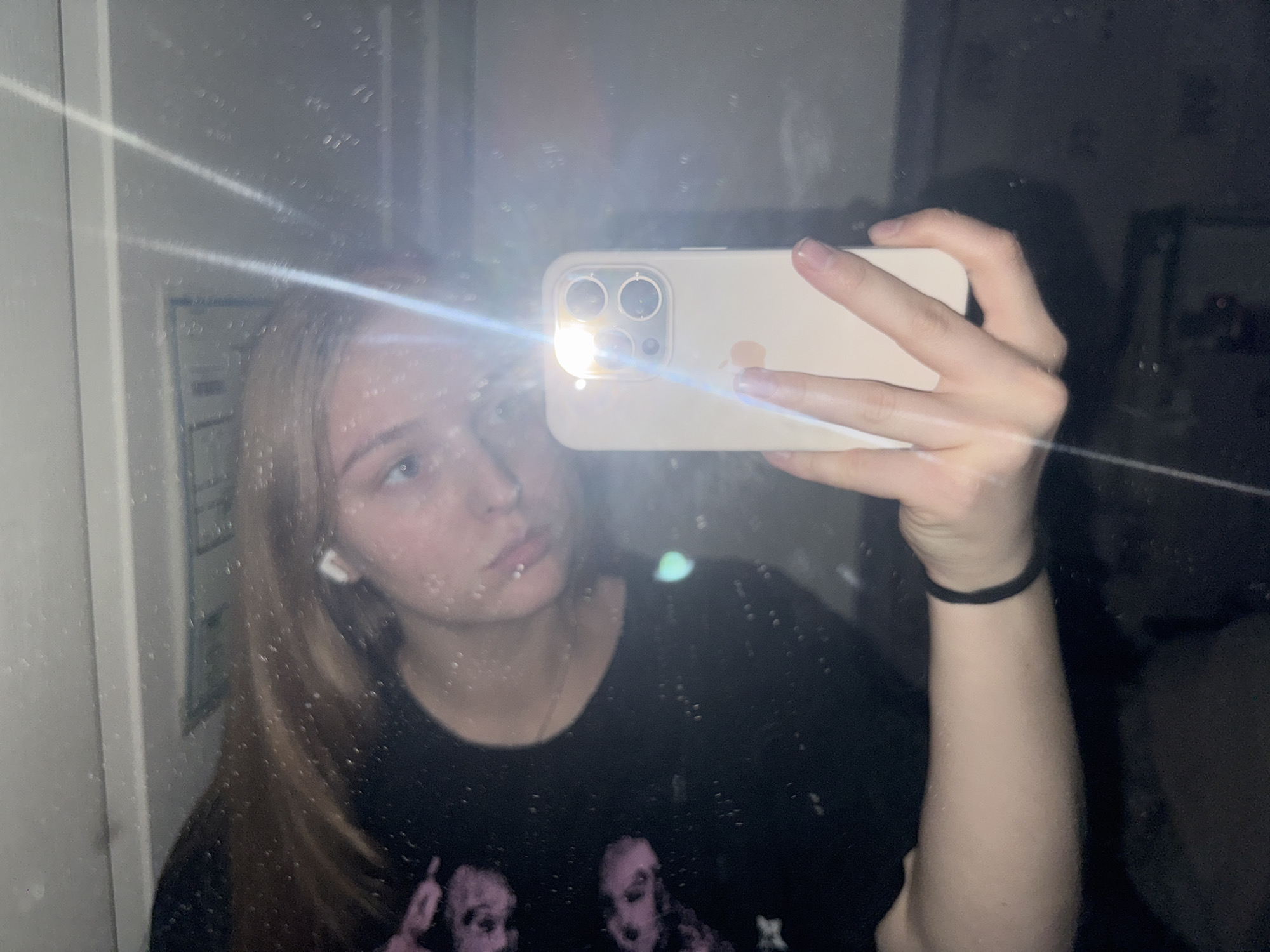 A young woman is taking a selfie in a mirror. She has blonde hair and is wearing a black t-shirt with a pink design. She is holding a light-colored smartphone, which is reflecting the flash. The mirror is dirty, with water spots and streaks, suggesting it might be in a bathroom or a similar environment. The background is dark and indistinct, with some blurry reflections that do not provide specific details about the location. There is no information in the image to indicate the location is in the city of Perm, Russia.
