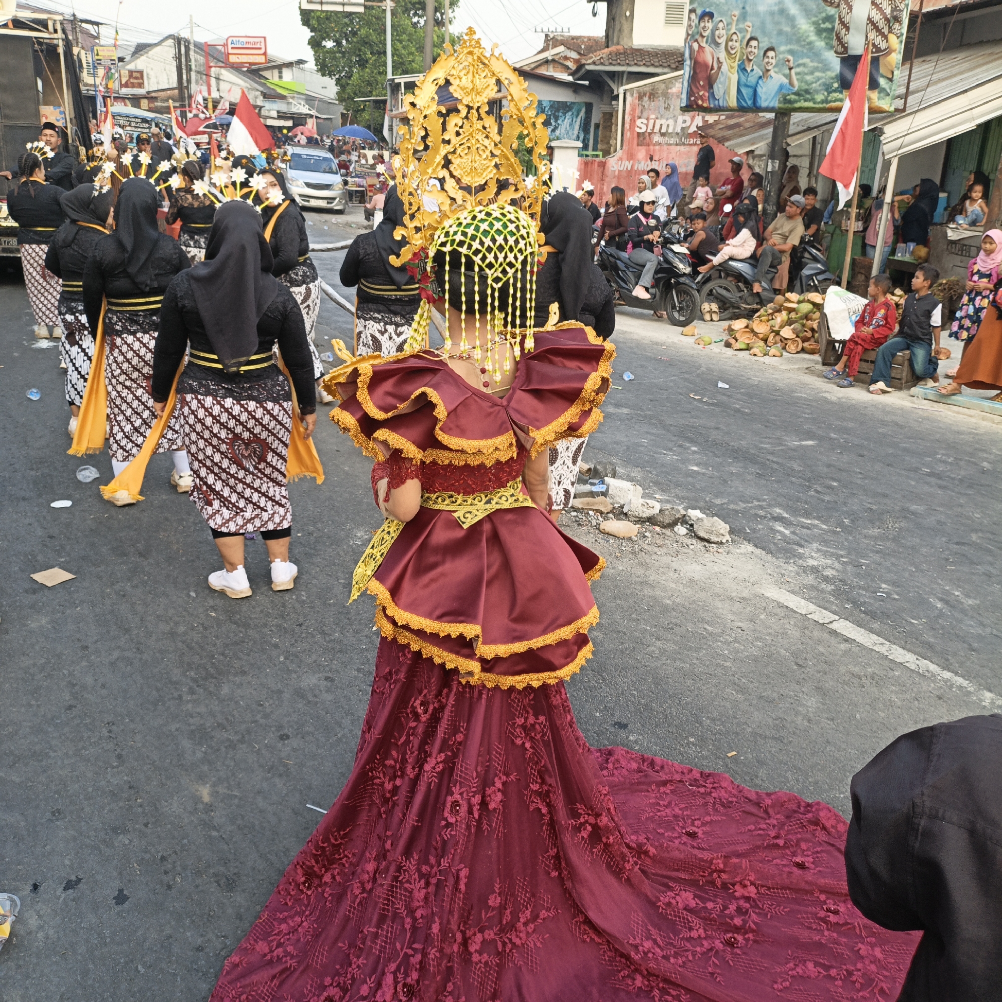 In Randudongkal, Indonesia, a procession is underway on a street lined with buildings and onlookers. At the forefront, a young person is adorned in a striking, elaborate maroon and gold traditional costume, complete with a tall, ornate golden headdress featuring intricate patterns and hanging beadwork. Their costume includes a ruffled, off-the-shoulder top with gold trim and a voluminous, floor-length skirt embellished with detailed floral embroidery.
Behind this central figure, several individuals in matching black tops, headscarves, and patterned batik skirts walk in formation, some holding yellow sashes. The procession is taking place on an asphalt road, with debris and litter scattered on the ground, including discarded coconut shells and plastic bottles.
The background reveals a bustling street scene with various vehicles, including motorcycles and cars. On the sides of the street, spectators are gathered, some sitting on curbs or on motorcycles, observing the event. Buildings line the street, some displaying advertisements and flags, including a red and white Indonesian flag. The overall atmosphere suggests a cultural festival or parade taking place in a residential or commercial area of the town.