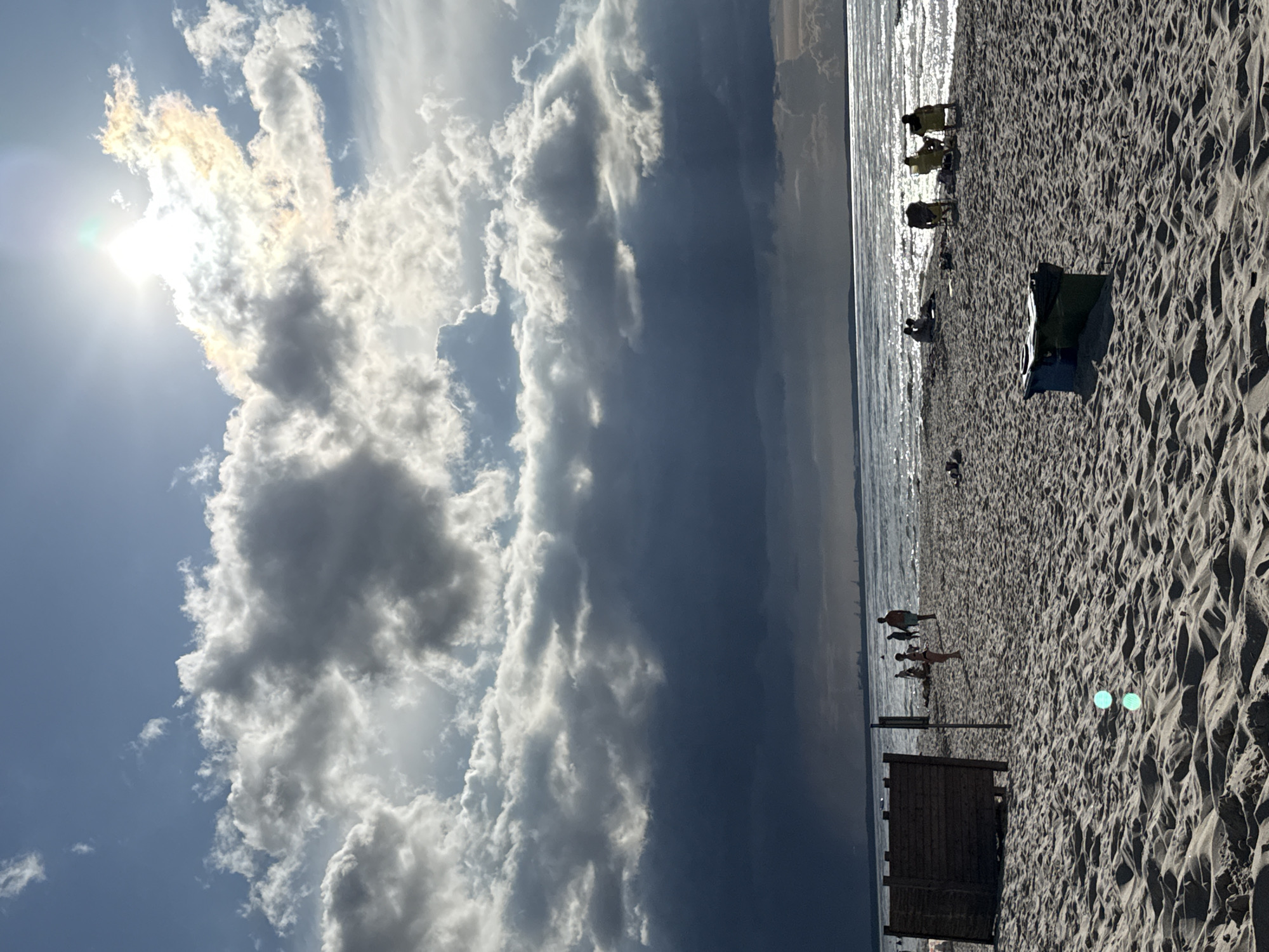 This is a photograph of a beach scene in Międzyzdroje, Poland. The sky is partly cloudy with dramatic dark clouds in the distance contrasting with brighter clouds closer to the sun, which is high and bright, causing a lens flare. The sun reflects off the water, creating a shimmering effect.
The beach is covered in sand with visible footprints and ripples. In the foreground, there are two bright turquoise circular artifacts. Further back on the sand, a dark wooden changing station or structure stands on the left. To the right of it, several people are visible, some walking and some standing.
Closer to the water's edge, a group of people are seated in chairs, some facing the water. Further in the background, the sea stretches to the horizon, where a faint outline of industrial structures or a distant city can be seen. The overall impression is a dynamic sky over a populated beach during daylight.