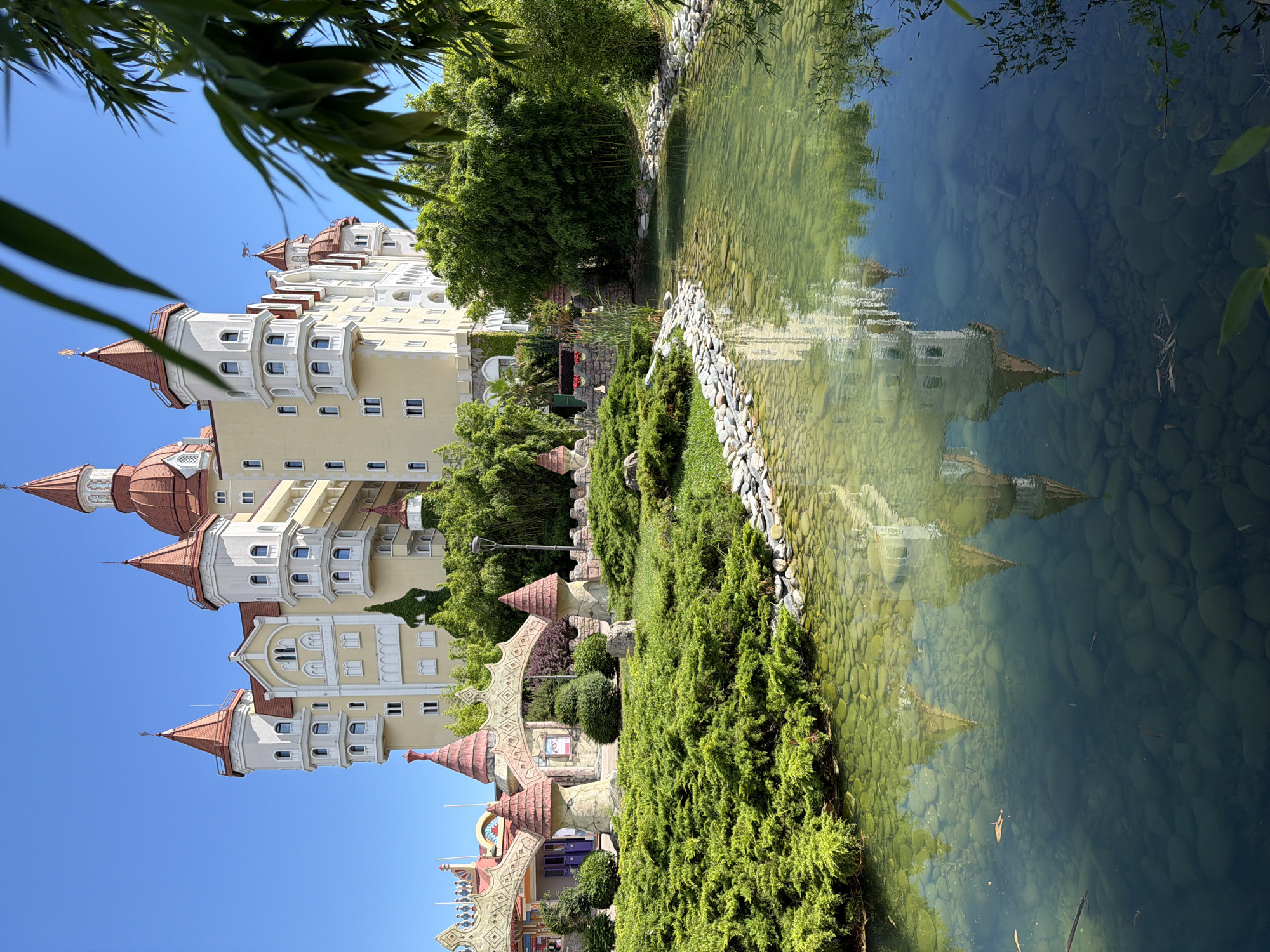 The image depicts a charming, fairy-tale-like hotel or resort building with a distinct castle-like architecture, featuring multiple turrets with reddish-brown conical roofs and cream-colored walls. The building has numerous windows and decorative elements. It is situated amidst lush greenery, including manicured bushes and trees, and is bordered by a clear, shallow pond. The pond's surface reflects the building and the surrounding foliage, showcasing a serene and picturesque scene.
The pond bed is visible, covered with smooth, rounded stones of varying sizes and colors, and the water appears calm and inviting. Several small fish are swimming in the pond. The sky is a clear, bright blue, suggesting a sunny day. The foliage appears vibrant and healthy, indicating a pleasant climate. The overall impression is one of tranquility and perhaps a tourist destination.
The image is oriented vertically, as if the viewer is looking up at the building from a low angle, or the photograph was taken on its side. The perspective of the castle structure, combined with the reflective pond and manicured gardens, creates a whimsical and idyllic atmosphere. The presence of a decorative arched gateway in the foreground further enhances the fairy-tale ambiance. No people are visible in the image. The time of day appears to be daytime, likely late morning or early afternoon, given the brightness of the sunlight. No visible text is present in the image. The location context provided is Sochi, Russia, which aligns with a resort setting that might feature such architecture and landscaping.