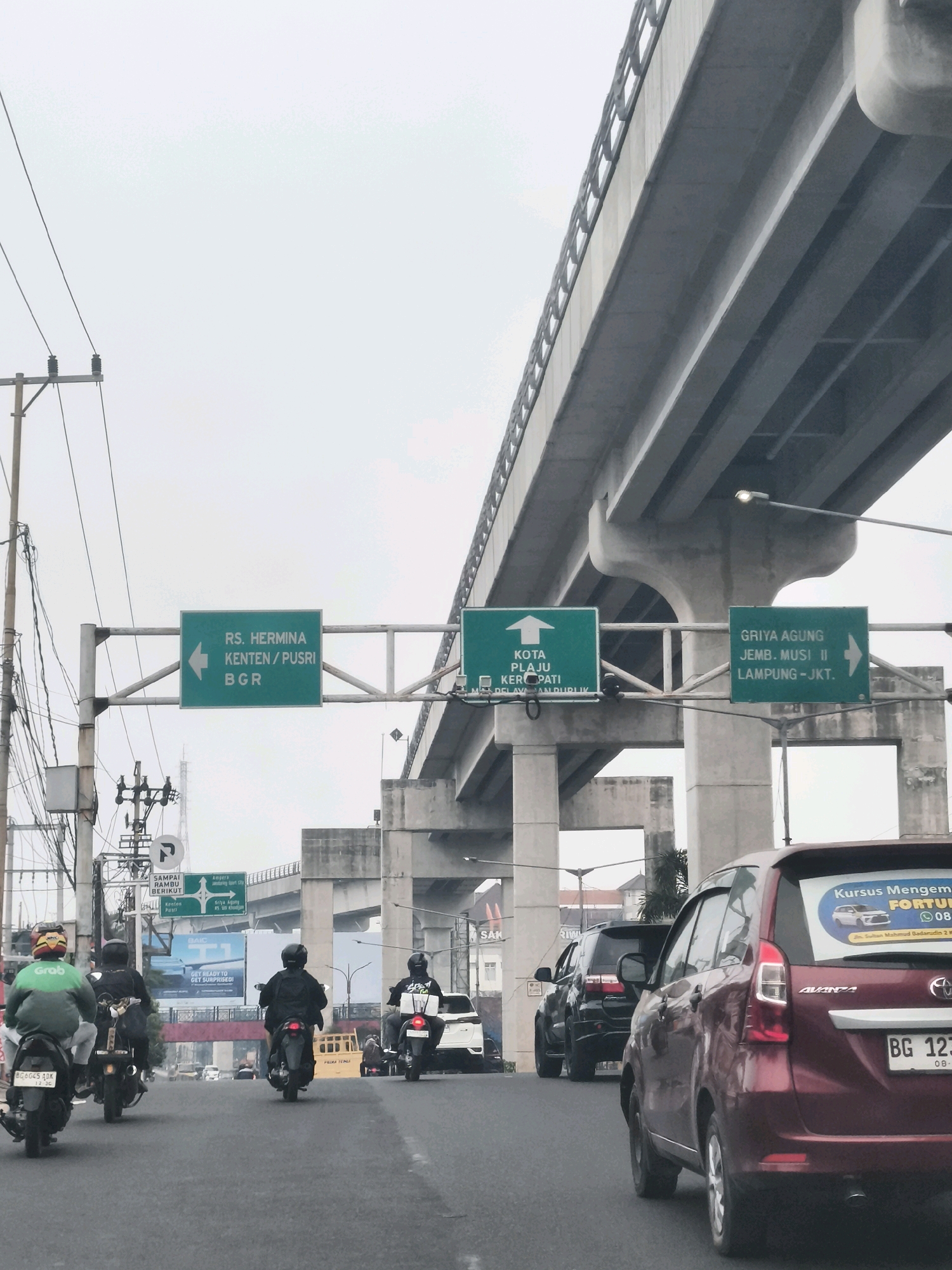 The image depicts a street scene in Palembang, Indonesia, likely on a cloudy day. A multi-lane road runs beneath an elevated concrete highway structure. Traffic consists primarily of motorcycles and cars. Several motorcyclists are visible in the foreground, some wearing helmets with logos like "Grab". Cars, including a maroon Toyota Avanza with a license plate starting with "BG 123" and a white SUV, are also present. Overhead signs provide directions to various locations, including "RS. Hermina Kenten/Pusri BGR", "Kota Plaju Kertapati", and "Griya Agung Jemb. Musi II Lampung-Jkt". Electrical poles and wires are prominent along the side of the road, and a billboard with text including "GET READY TO GET SURPRISED!" is visible in the mid-ground. The overall atmosphere is that of urban traffic.