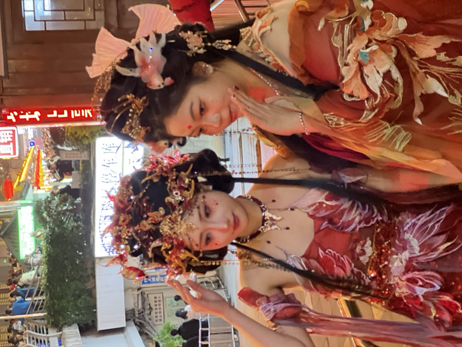 Two East Asian females, dressed in elaborate traditional Chinese-style attire, are depicted in a medium close-up at night in a brightly illuminated urban setting within Yuzhong District, China. The subject on the left wears a sleeveless red gown featuring intricate white and red embroidery, sequin embellishments, and sheer flowing elements. Her hair is styled in an ornate black updo adorned with gold and red phoenix-like headpieces, dangling jewels, and a forehead ornament. She also wears a dark red choker with a silver-toned winged pendant. Her left hand is raised, fingers delicately touching her left temple near her headpiece. The subject on the right is dressed in a layered orange and red gown with broad sleeves, detailed with gold, orange, and teal floral embroidery. Her hair is styled in a similar dark updo, embellished with gold ornaments, a large white fan-shaped piece, and two illuminated pinkish-white fish ornaments. Her right hand is raised, with fingers touching her chin/cheek area. Both women display elaborate facial makeup, including glitter and a red mark between their eyebrows. The background reveals a multi-level street environment, brightly lit by warm artificial lights and red neon signs. Visible elements include a metal railing, green foliage, a partial white sign with Chinese characters translating to "Parking Garage," and a vertical red sign with characters translating to "Direct Sale." Numerous casually dressed pedestrians are observable on an elevated walkway and at street level in the blurred background, indicative of a bustling public space. A wooden wall and window frame are visible to the far right.