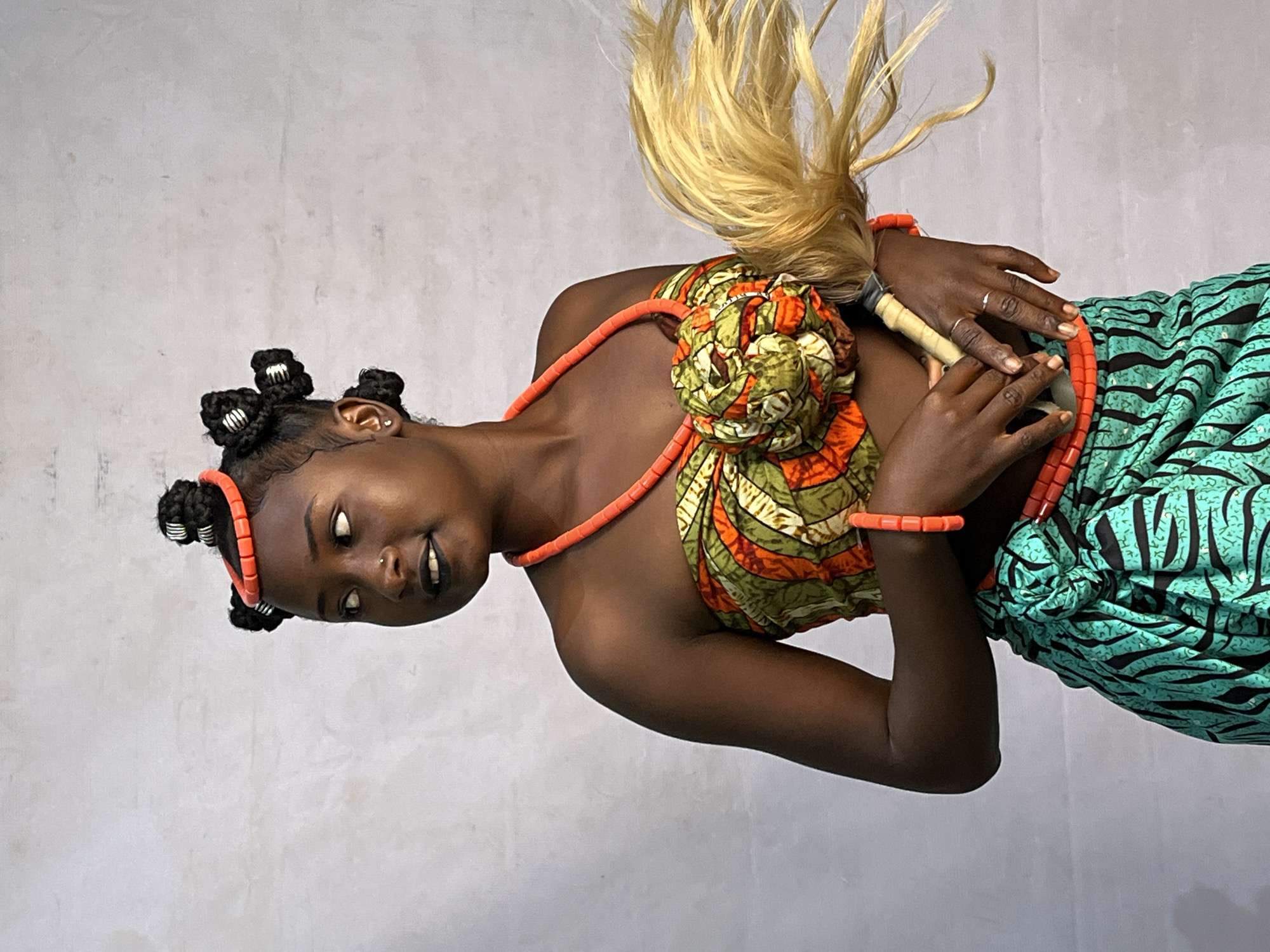 In a medium shot, a young woman with dark skin stands against a plain, light-colored, textured wall. She is adorned in traditional Nigerian attire, featuring a brightly colored, patterned strapless top made of fabric with orange, green, and white designs, cinched at the bust with a decorative knot. Her lower garment is a teal skirt with a black zebra-like pattern, tied at the waist. She wears multiple strands of coral-colored beads around her neck and wrists, and a single matching bracelet on her right arm. Her hair is styled in multiple black buns, some of which are encircled with coral beads. Her makeup includes dark lipstick and a subtle nose ring. In her hands, she holds a whisk-like object made of light-colored straw or hair, with the handle visible. The woman looks to her left, her head tilted slightly, with a gentle smile. The lighting suggests an indoor setting, possibly a studio, with the backdrop being the wall of a building in Barakin Akawo, Nigeria.