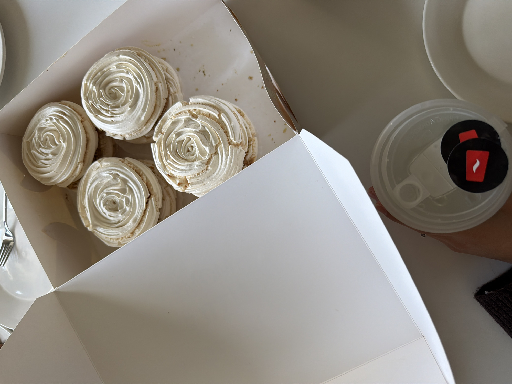 A high-angle shot depicts an open white box containing five meringues, characterized by a swirled cream topping. Adjacent to the box, a clear plastic cup with two circular black stickers bearing a stylized red flame design is held by a person. The scene unfolds in Yerevan, Armenia, evidenced by the logo on the plastic cup. Cutlery and plates are also partially visible on the table.