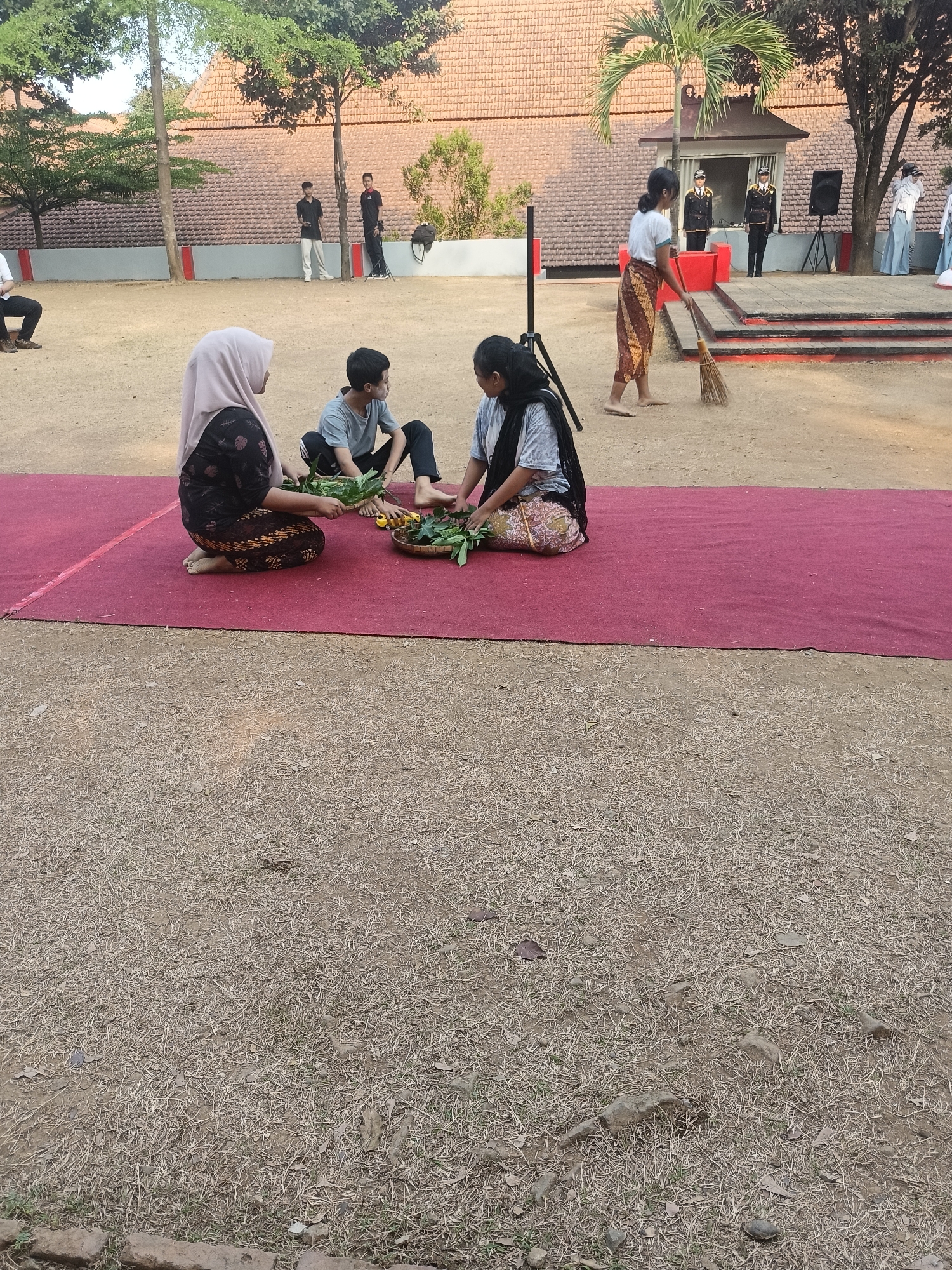 In Pengkol, Indonesia, three individuals are seated on a maroon carpet laid on dry, dusty ground, engaged in an activity involving a woven basket filled with greenery and yellow objects. To the left, a woman wearing a pink hijab and a dark patterned dress is partly visible. In the center, a young person in a grey t-shirt and black pants is seated. To the right, a person in a dark shirt and patterned skirt also participates in the activity. In the background, a stage area is visible with two individuals in uniform standing behind a desk. A woman to the right of the stage is sweeping the ground with a broom. Other figures in uniform are also present in the background. Trees and buildings with tiled roofs are part of the outdoor environment.