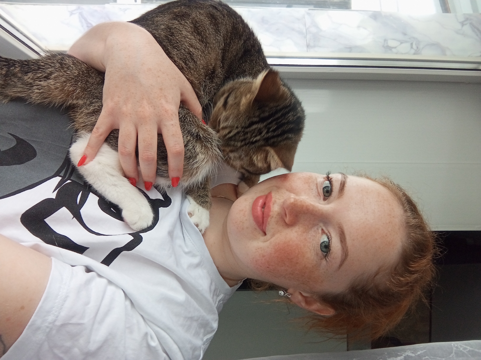 A young, light-skinned woman with freckles and red nail polish is holding a tabby cat. She has red hair and blue eyes, and is wearing a white t-shirt with a black design. The cat is resting on her chest. The background suggests an interior setting, possibly near a window with a marble-like sill. There is no information to indicate this image was taken in Yurgamysh, Russia.