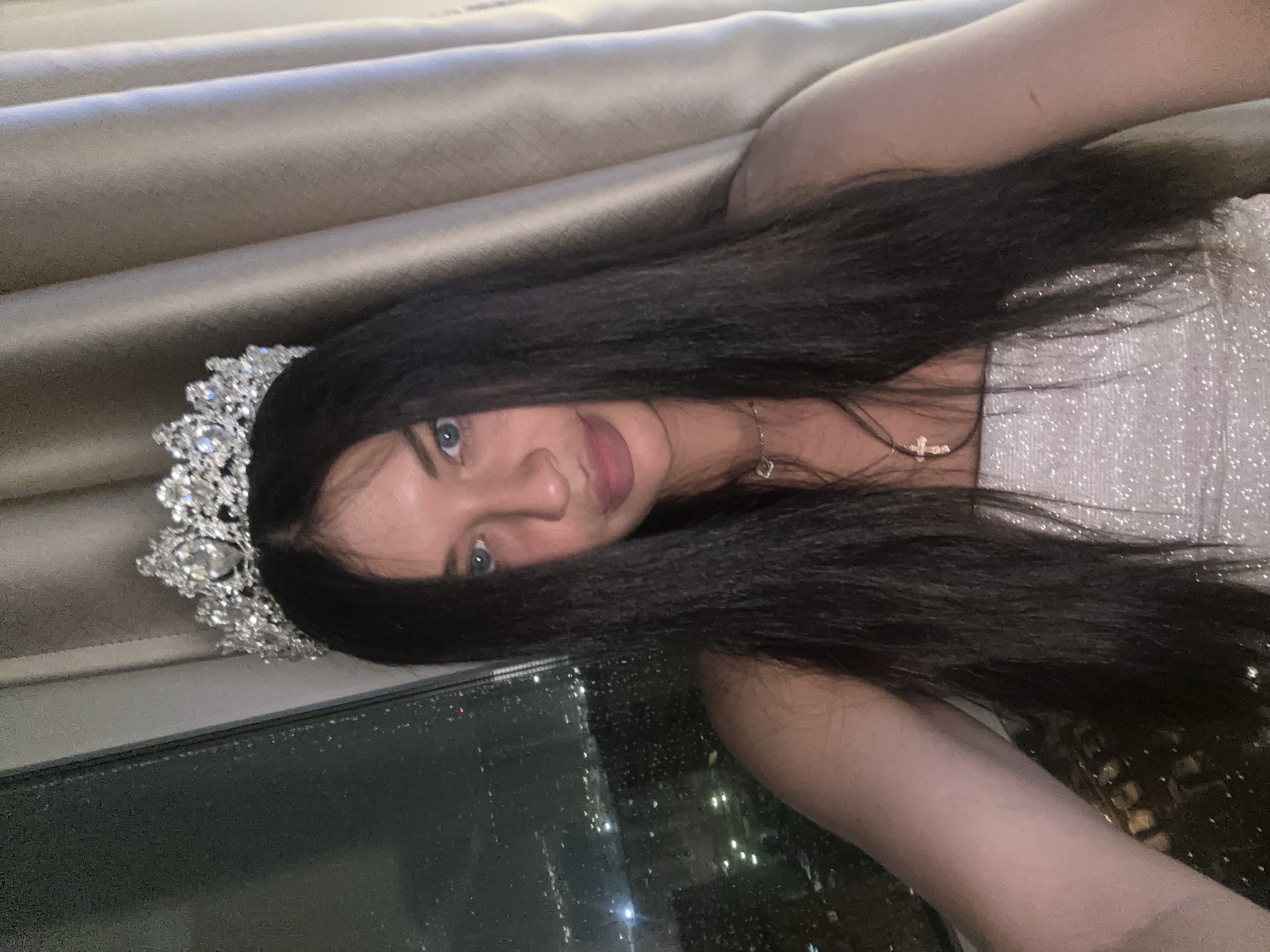 A young woman is captured in a close-up, slightly overhead shot, lying down and looking directly at the camera with a gentle smile. She has long, dark hair fanned around her and striking blue eyes. A detailed, ornate silver tiara adorns her head. She wears a sparkly, light-colored strapless top or dress and two delicate necklaces: one featuring a small silver cross pendant and another with a heart-shaped pendant.
The scene is set indoors, likely in a high-rise building like a hotel room or apartment, offering a panoramic view. The upper background features beige or light-brown pleated curtains made of a smooth fabric. Below her, a large window showcases a nighttime city skyline, brightly lit with countless building and street lights. Visible water droplets on the window glass suggest rain or condensation outside. The overall lighting is soft, highlighting the woman while allowing the illuminated cityscape of Moscow, Russia, to serve as a dramatic backdrop. The time of day is clearly night. The woman appears calm and relaxed. No visible text is present in the image.