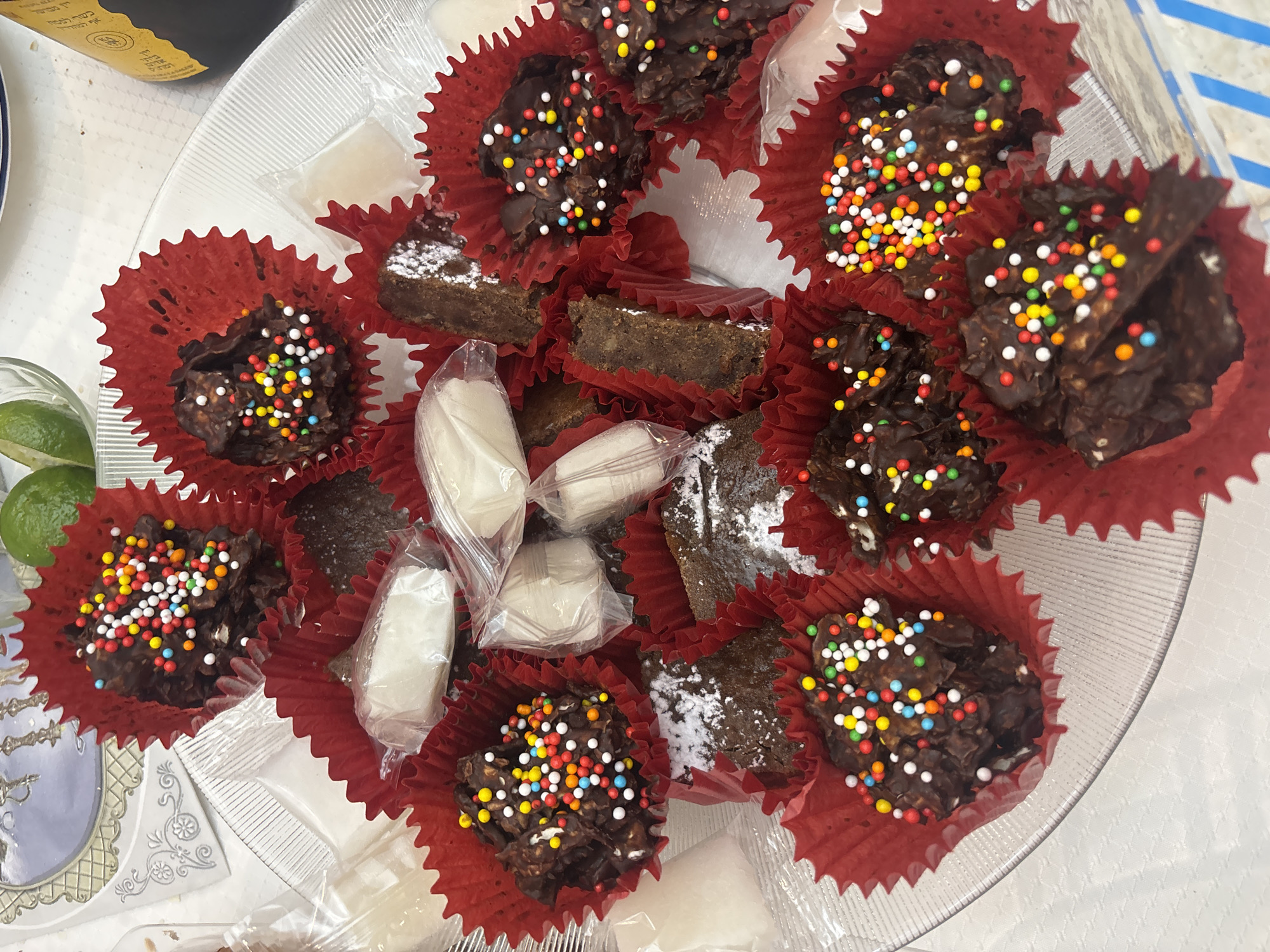 A clear glass plate filled with an assortment of sweets in red paper cupcake liners is displayed. Some of the treats are chocolate clusters with colorful sprinkles, while others appear to be brownies, some dusted with powdered sugar. There are also individually wrapped white candies scattered among the other sweets. A partial view of green limes and a bottle can be seen in the background. The location of this scene is Huixquilucan de Degollado, Mexico.