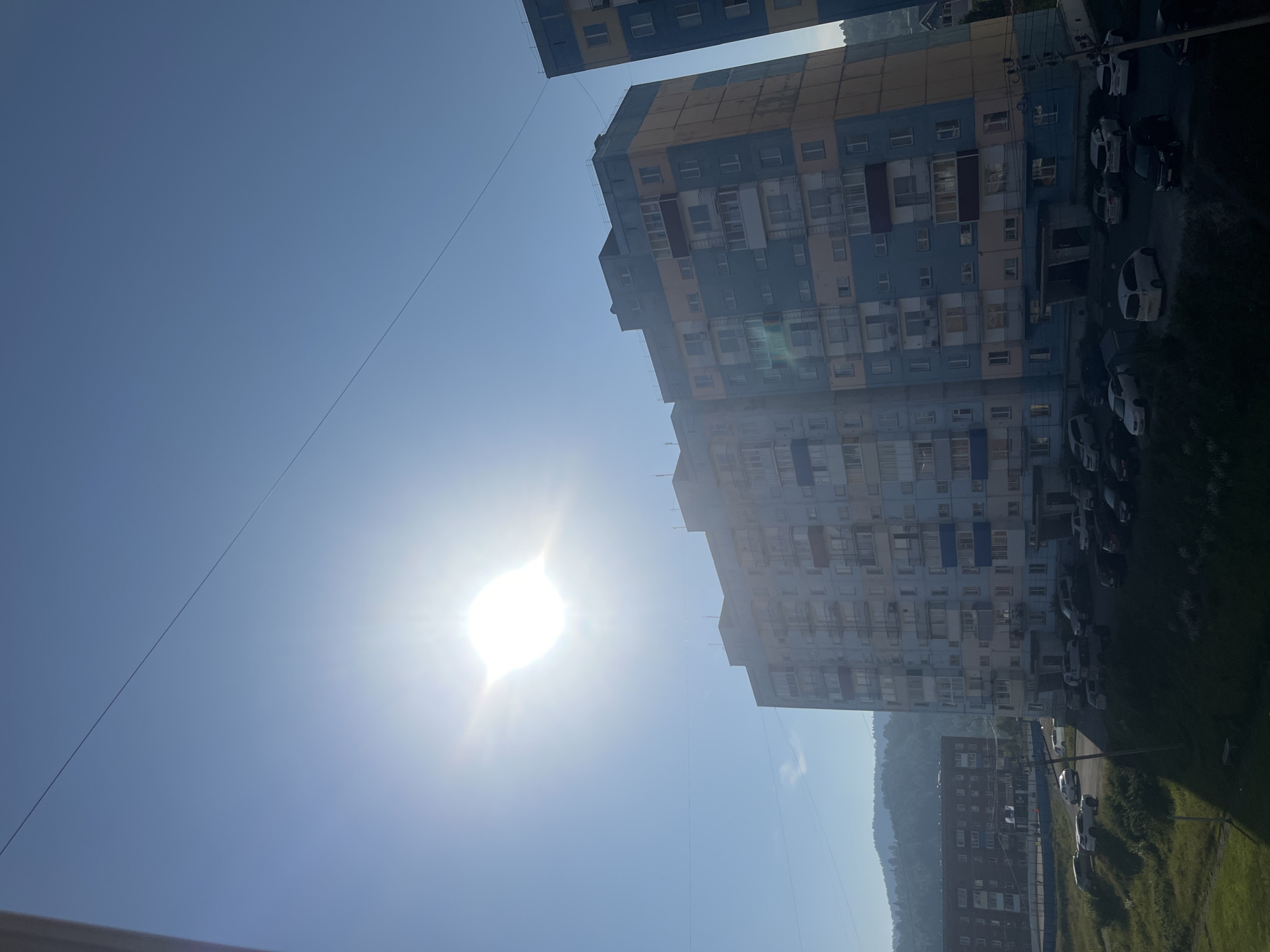 This is a photograph taken in Sheregesh, Russia, depicting a residential apartment building under a clear blue sky with the sun prominently visible and creating a lens flare. The building is multi-story, with a facade painted in a combination of light blue and yellow. Numerous balconies are visible on the building's exterior. In front of the building, a parking area contains several cars of various makes and models. To the left, another apartment building is partially visible. In the background, rolling hills with a light haze are present, suggesting a mountainous or hilly terrain. A thin wire runs diagonally across the sky. The overall scene is a daytime view of a residential area.