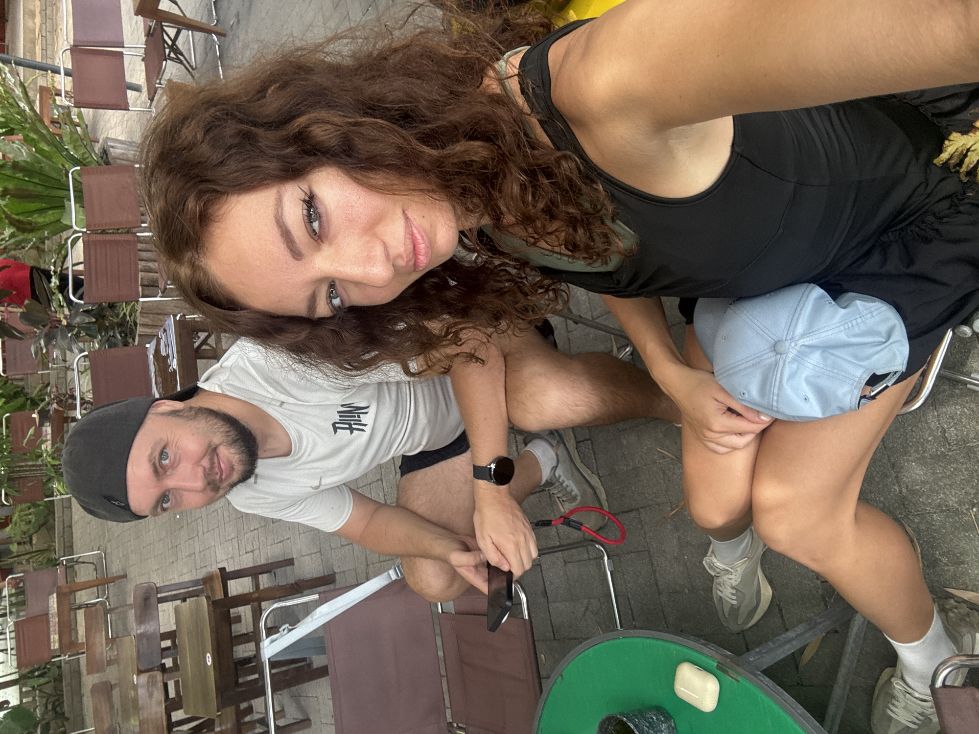 A photograph captures two individuals posing for a selfie in an outdoor setting. A woman with curly brown hair, wearing a black tank top and shorts, holds the camera and looks directly at it. Next to her, a man with a beard and a black baseball cap is also looking towards the camera while holding a phone in his hand. They are sitting at tables surrounded by empty brown folding chairs, likely at a cafe or restaurant. A green table with some items on it is in the foreground. The location is Đà Nẵng, Vietnam, based on the general appearance and foliage in the background.