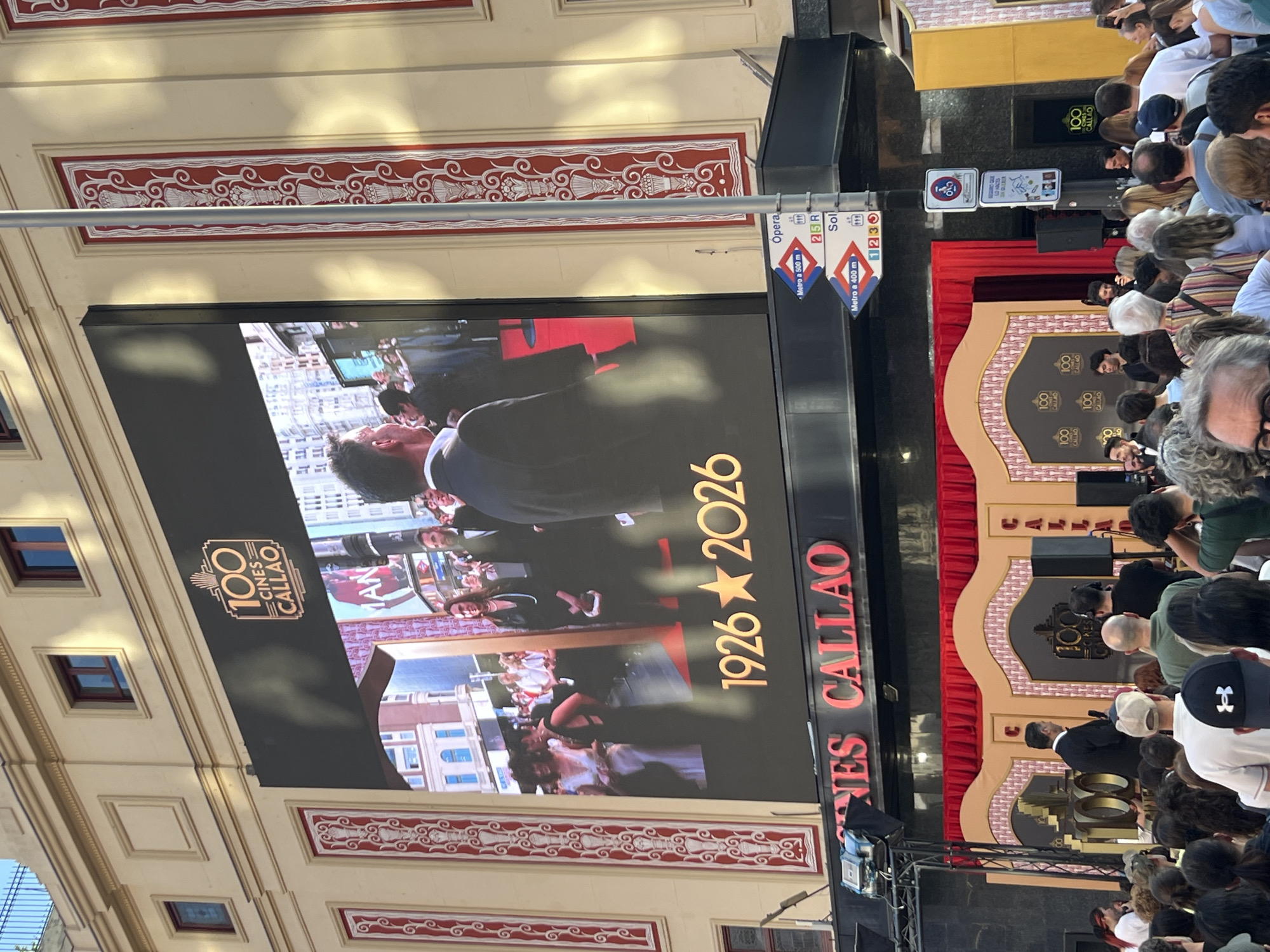 The image captures an outdoor event in Madrid, Spain, likely related to the 100th anniversary of the Cines Callao. A large outdoor screen displays images associated with the anniversary, along with the dates "1926-2026" and the text "CINES CALLAO". In front of the screen, a crowd of people has gathered, suggesting a public event or presentation. There is a stage with a backdrop, possibly for speakers or performers. Signage for the Opera and Sol metro stations is also visible. The building facade, adorned with decorative elements, adds to the urban setting of the event. The image conveys a sense of festivity and public engagement surrounding this centennial celebration.