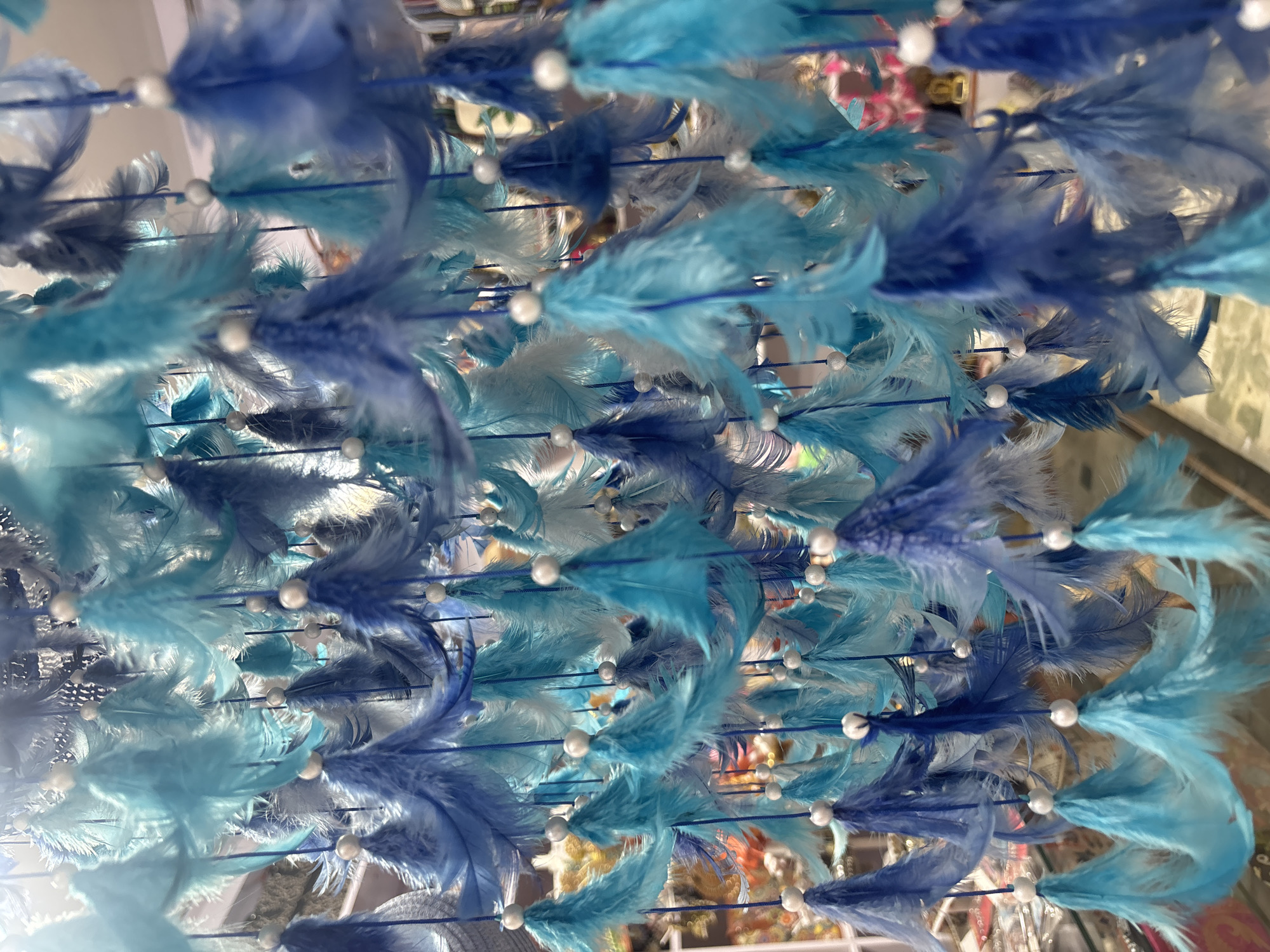 A close-up, eye-level shot showcases a hanging decoration composed of numerous blue and teal feathers and white beads. The feathers are arranged in clusters, with each cluster attached to a thin blue string. The strings are suspended from above, creating a cascading effect. The background is blurred, with hints of other decorations or merchandise visible, suggesting a marketplace or shop setting. The lighting is bright, causing some specular highlights on the beads and feathers. The overall impression is of a vibrantly colored, textured object, likely a dreamcatcher or a similar decorative item, possibly found in the bustling markets of Vashisht, India, known for its spiritual retreats and souvenir shops.