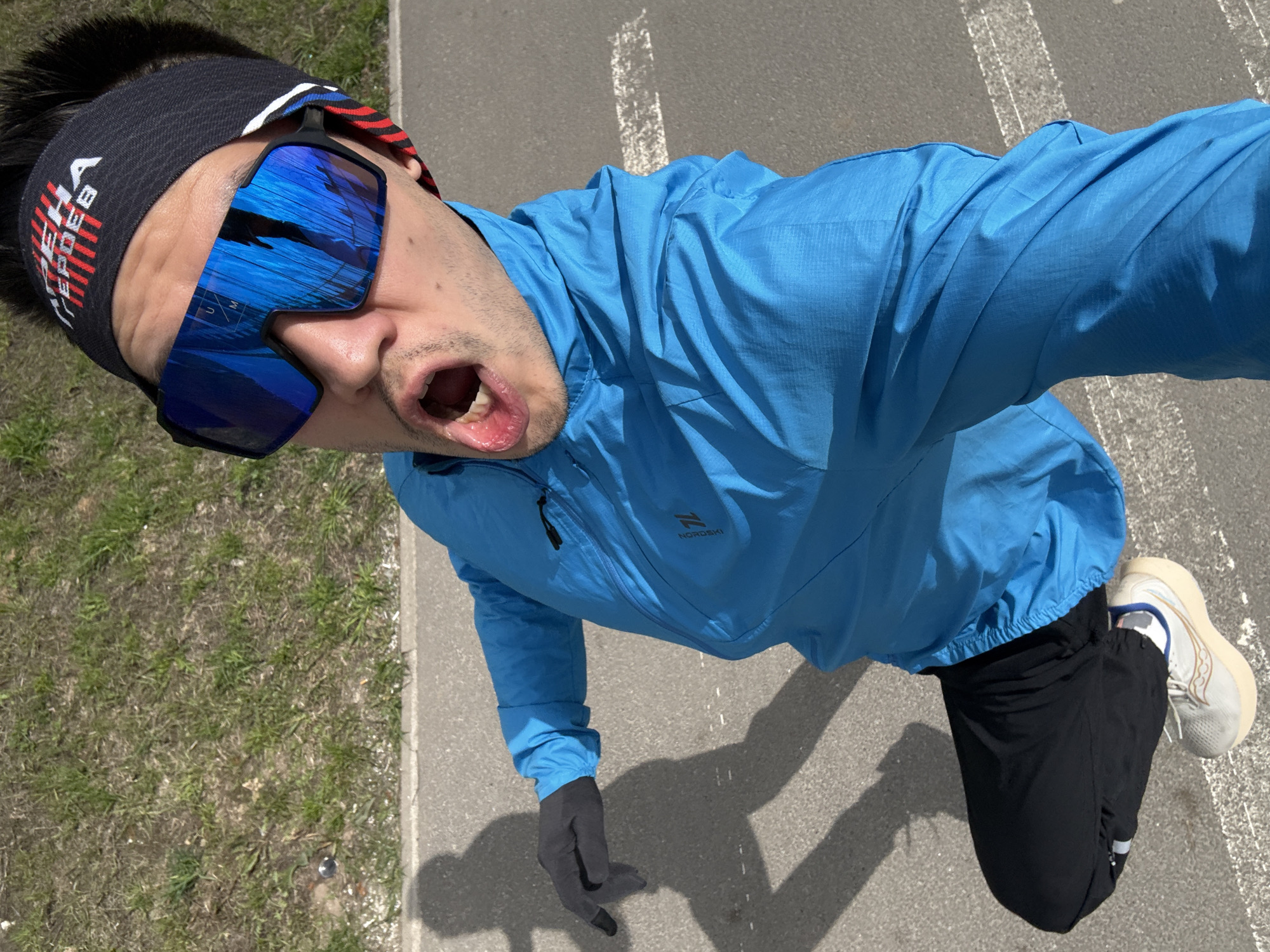 A high-angle selfie captures a male-presenting individual from above, portraying him engaged in an outdoor activity, likely in Kazan, Russia, given the context. The person is dressed in athletic gear, including a bright blue lightweight jacket with a "NORD SKI" logo on the left chest, black athletic pants, and light-colored running shoes with white soles. He wears a black headband with red and white stripes, featuring the Russian text "ГОРДА ГЕРОЕВ" (Pride of Heroes), and large blue mirrored sports sunglasses that reflect the sky. His mouth is open, revealing teeth, suggesting exertion or a dynamic, enthusiastic pose. Dark grey gloves with black fingertips cover his hands.
The setting is an outdoor paved area, likely an asphalt path or road, bordered by a strip of dry, sparse grass on the left. Faint white markings are visible on the asphalt. The scene is brightly lit by strong sunlight, casting distinct shadows of the person onto the ground, indicating a clear, sunny day. The sunglasses also have a small "UM" visible on the bridge.