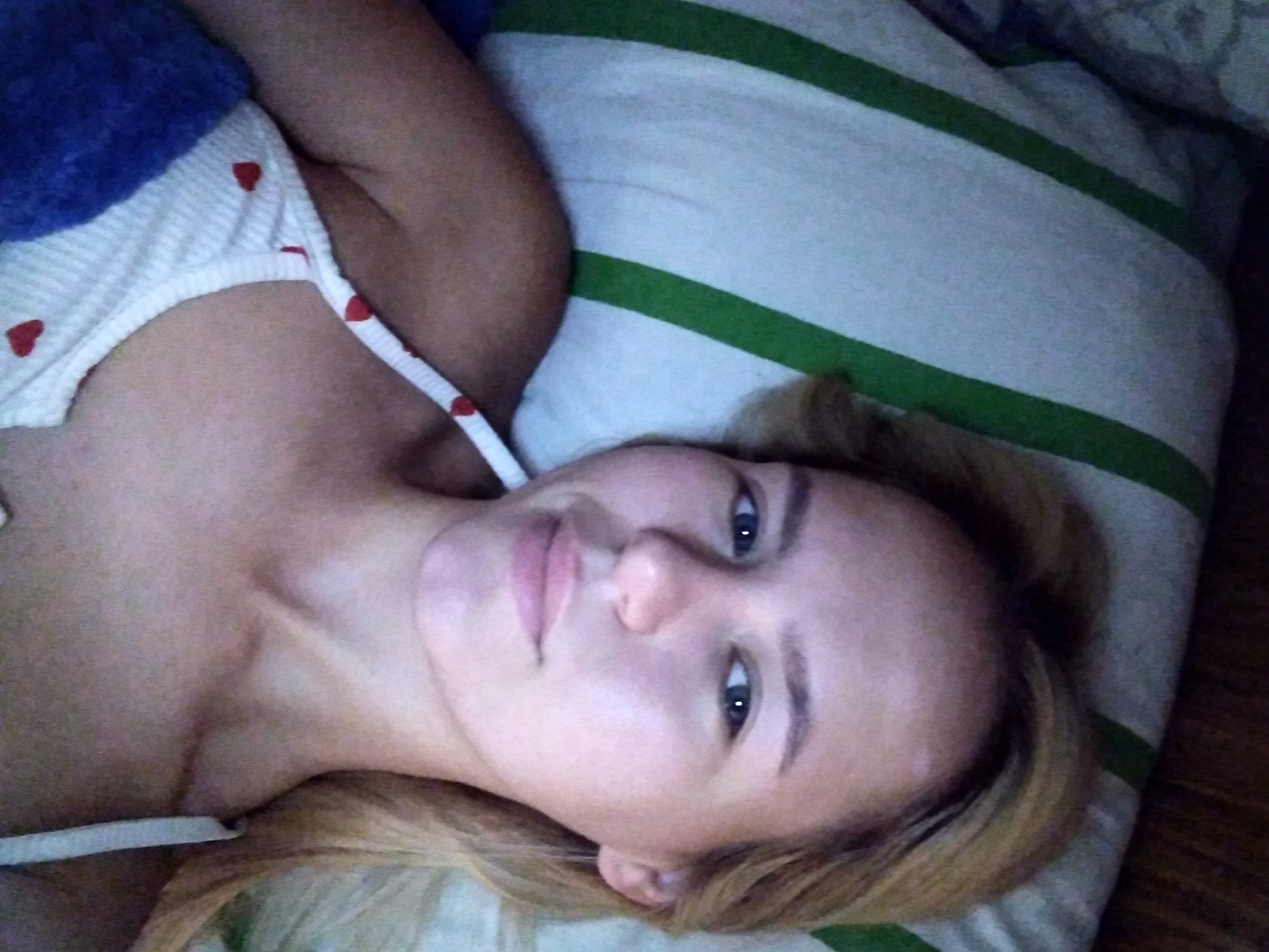 A young blonde woman is lying on her back in bed, looking at the camera. She is wearing a white tank top with small red hearts on it and a blue blanket is draped over her left shoulder. She has light skin and her hair is light brown. The background consists of a pillow with green and white stripes and a wooden headboard. The image appears to have been taken in a bedroom in Radisne, Ukraine.