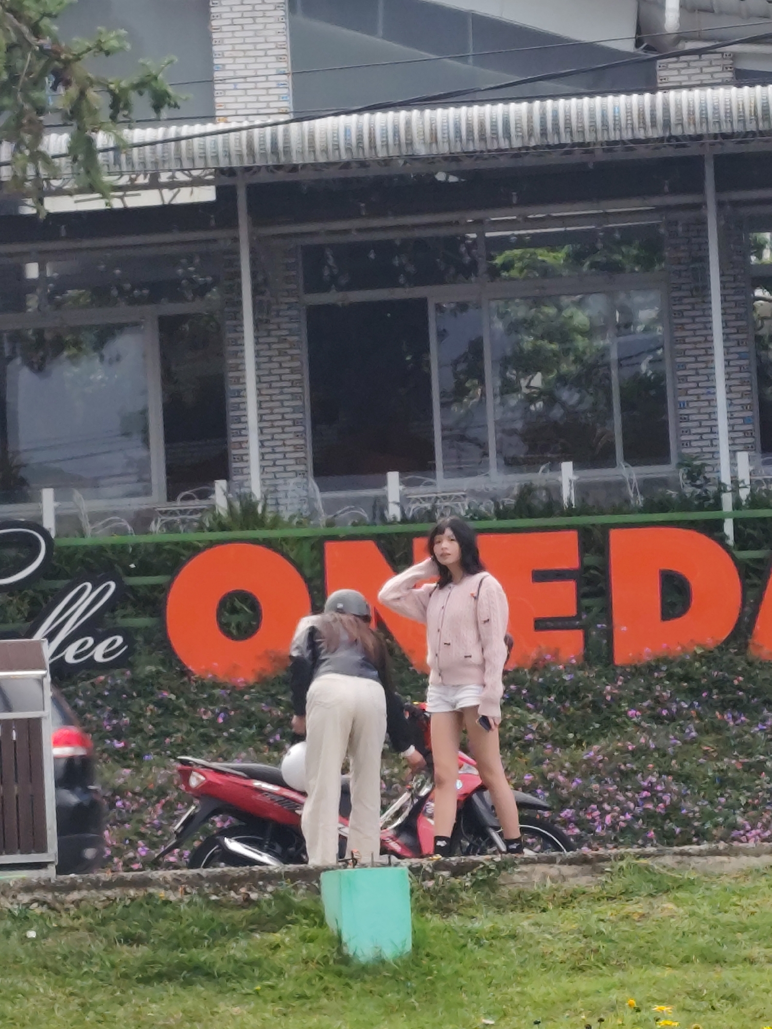 In Da Lat, Vietnam, two young women are captured in a street scene. The woman on the left, wearing a grey jacket and light-colored pants, stands near a red motorcycle. The woman on the right, dressed in a pink cardigan and white shorts, poses with her hand touching her hair and holding a phone. They are positioned in front of a large, orange sign that spells out "ONE DAY" in bold, block letters. The sign is set against a backdrop of lush green foliage and flowering plants. Behind them, a building with large windows and a striped awning suggests a commercial establishment, possibly a cafe or restaurant, given the visible text "Coffee" on the left side of the orange sign. The overall setting appears to be an outdoor area, with grassy ground in the foreground.