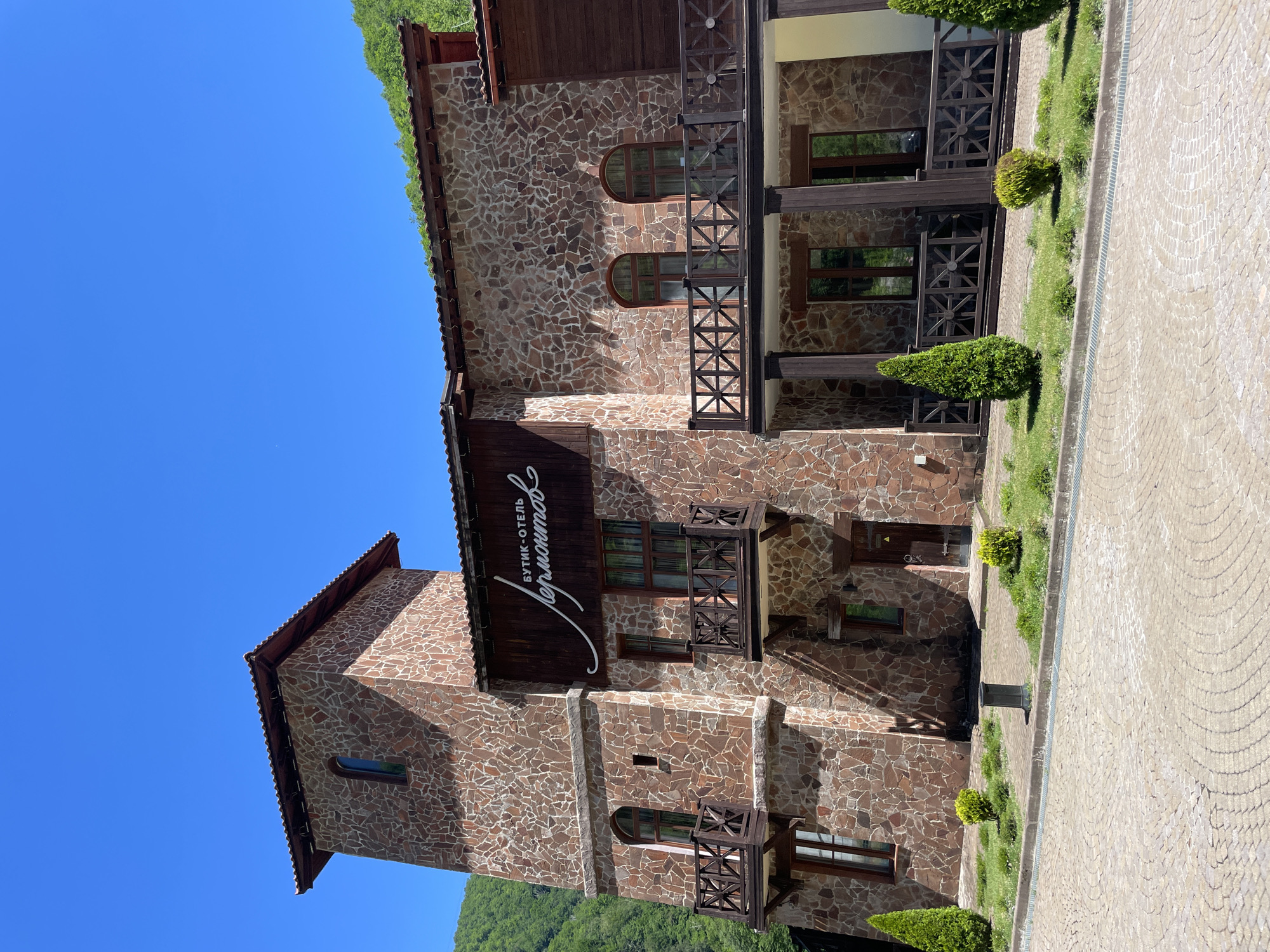 This is a daylight, eye-level, exterior shot of a boutique hotel named "Lyudmila" in the city of Estosadok, Russia. The hotel is constructed with rough, reddish-brown stone and dark wood accents. It features a tall, square tower with a single arched window and a pitched, red-tiled roof. The main building has multiple stories with balconies adorned with wooden railings and dark pillars. Several arched windows are visible, and a prominent wooden sign with the hotel's name in cursive script is mounted above the first floor. Small, manicured evergreen trees and bushes are planted in front of the hotel, and the ground is paved with light-colored, circular stone patterns. The background shows a steep, tree-covered hillside under a clear blue sky.