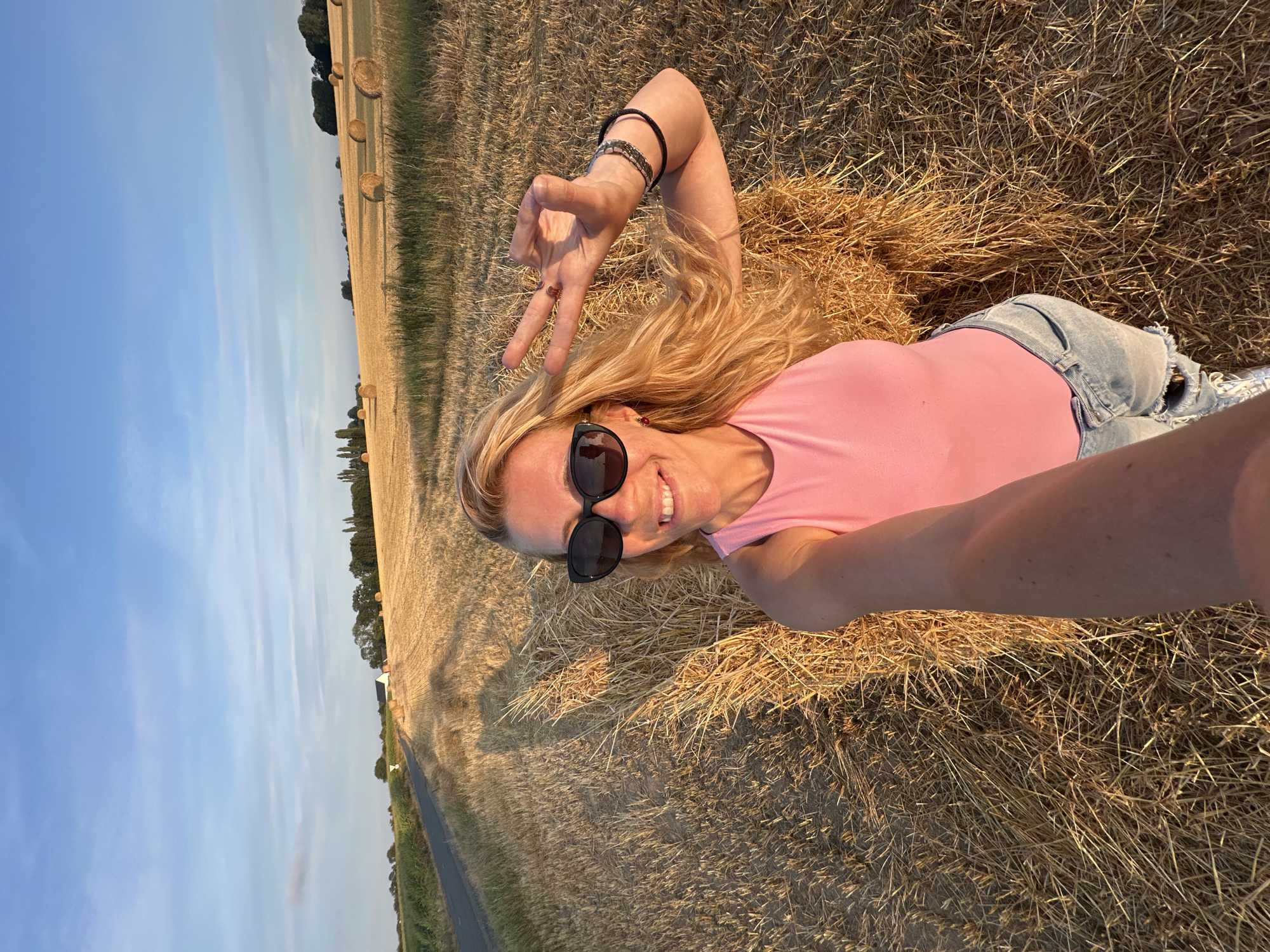 A blonde woman with sunglasses and a pink tank top is taking a selfie in a field of hay bales. She is smiling and holding up two fingers in a peace sign. The sun is setting, casting long shadows across the field. The location is specified as Trouville-Alliquerville, France, implying the image was taken in or around this area. The environment is rural, characterized by agricultural fields and a clear sky.