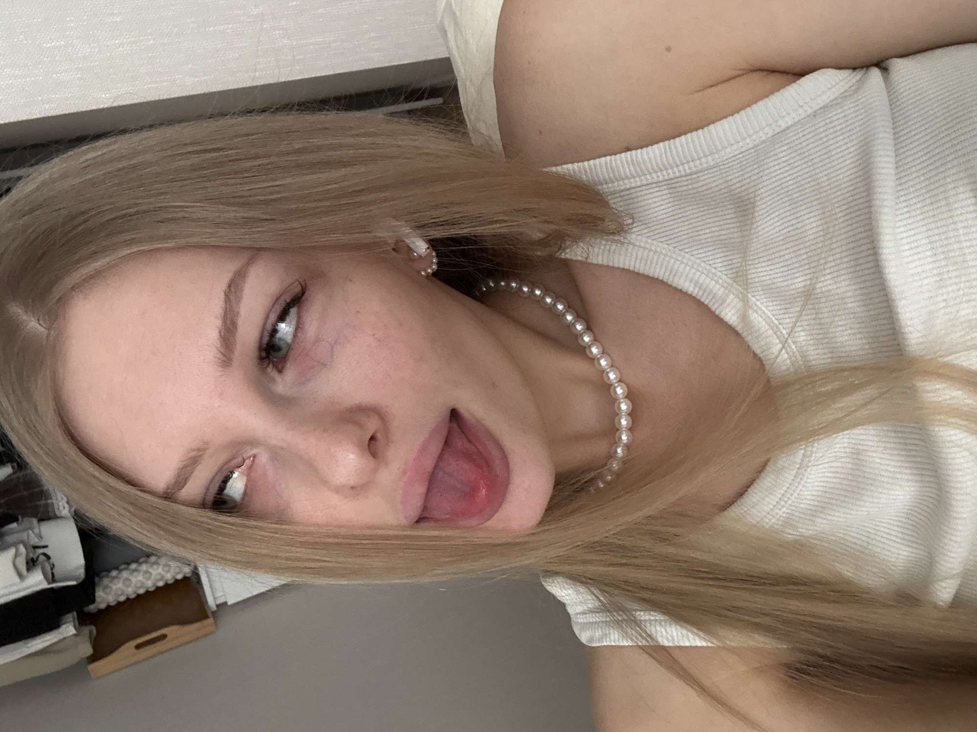 A woman with long blonde hair is lying down, possibly on a bed, wearing a white tank top and a pearl necklace. She has her tongue sticking out and is looking to her left. There's what appears to be a white headboard above her. An object near the lower left of the image seems to be a storage tray or box. There is no information to suggest that this image was taken in Perm, Russia.