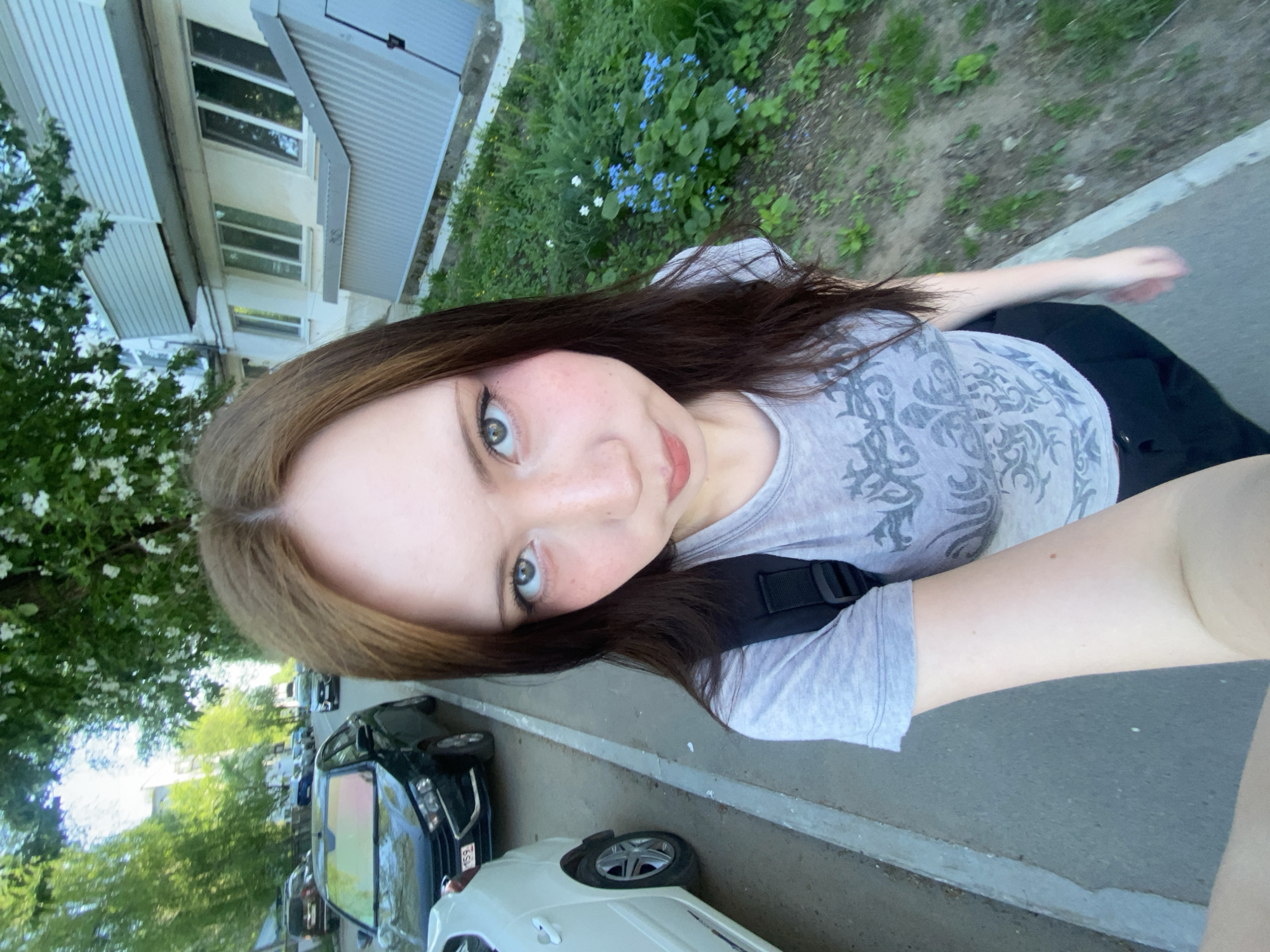 A young woman with brown hair, fair skin, and green eyes takes a selfie. She is wearing a gray t-shirt and a black backpack strap is visible over her shoulder. The background includes a multi-story building with windows, some greenery with flowers, a sidewalk, and parked cars. The location appears to be a city street.
